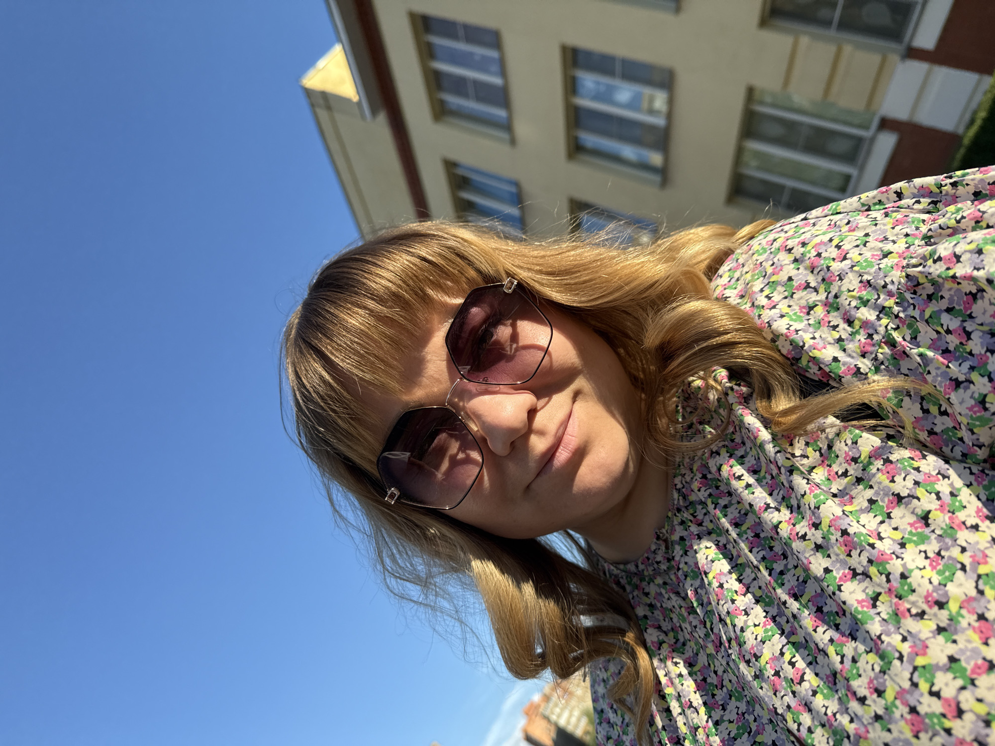 A young woman with blonde hair, wearing sunglasses and a floral dress, is pictured outdoors on a sunny day. She is looking up at the camera with a slight smile. The background shows a light-colored building with windows and a clear blue sky. The scene appears to be a candid shot, possibly a selfie, taken in daylight. There are no visible text elements in the image. The presence of a building suggests an urban or suburban setting. The bright sunlight indicates it is daytime. The woman's relaxed expression and attire suggest a casual and pleasant atmosphere.