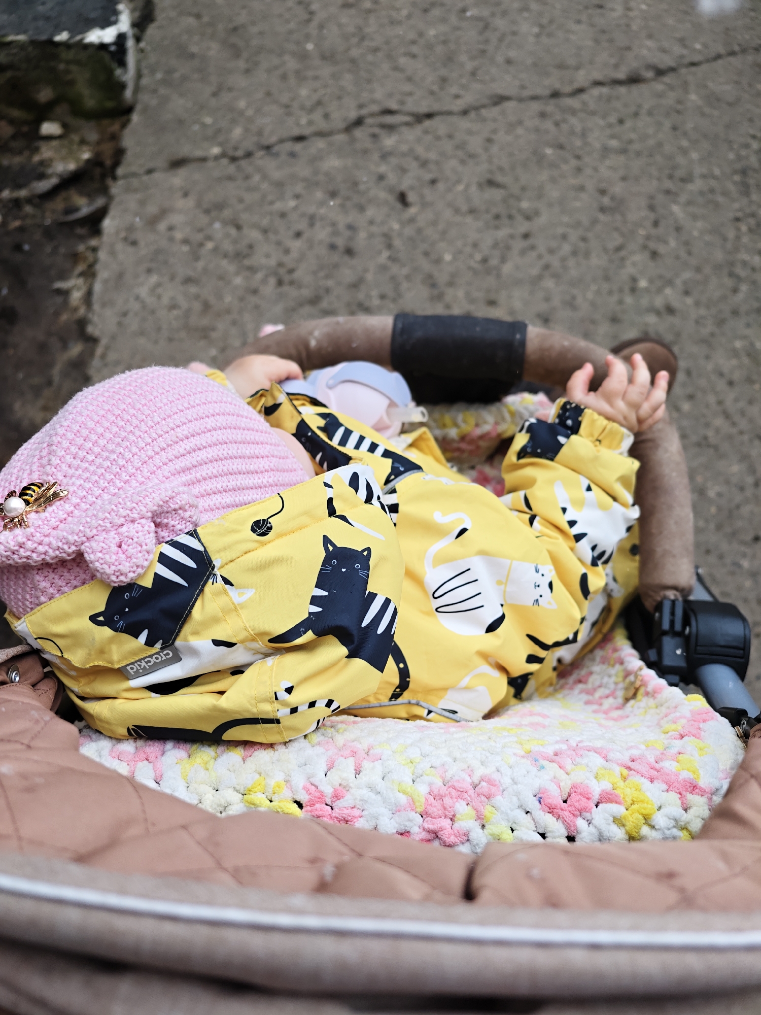 A baby wearing a yellow and black cat-print jacket and a pink hat with a bee-shaped pin is lying in a stroller. The baby is covered with a white, pink, and yellow blanket. The stroller is parked against a textured gray wall. The brand name "crockid" is visible on the jacket. The location is Chaikovsky, Russia.