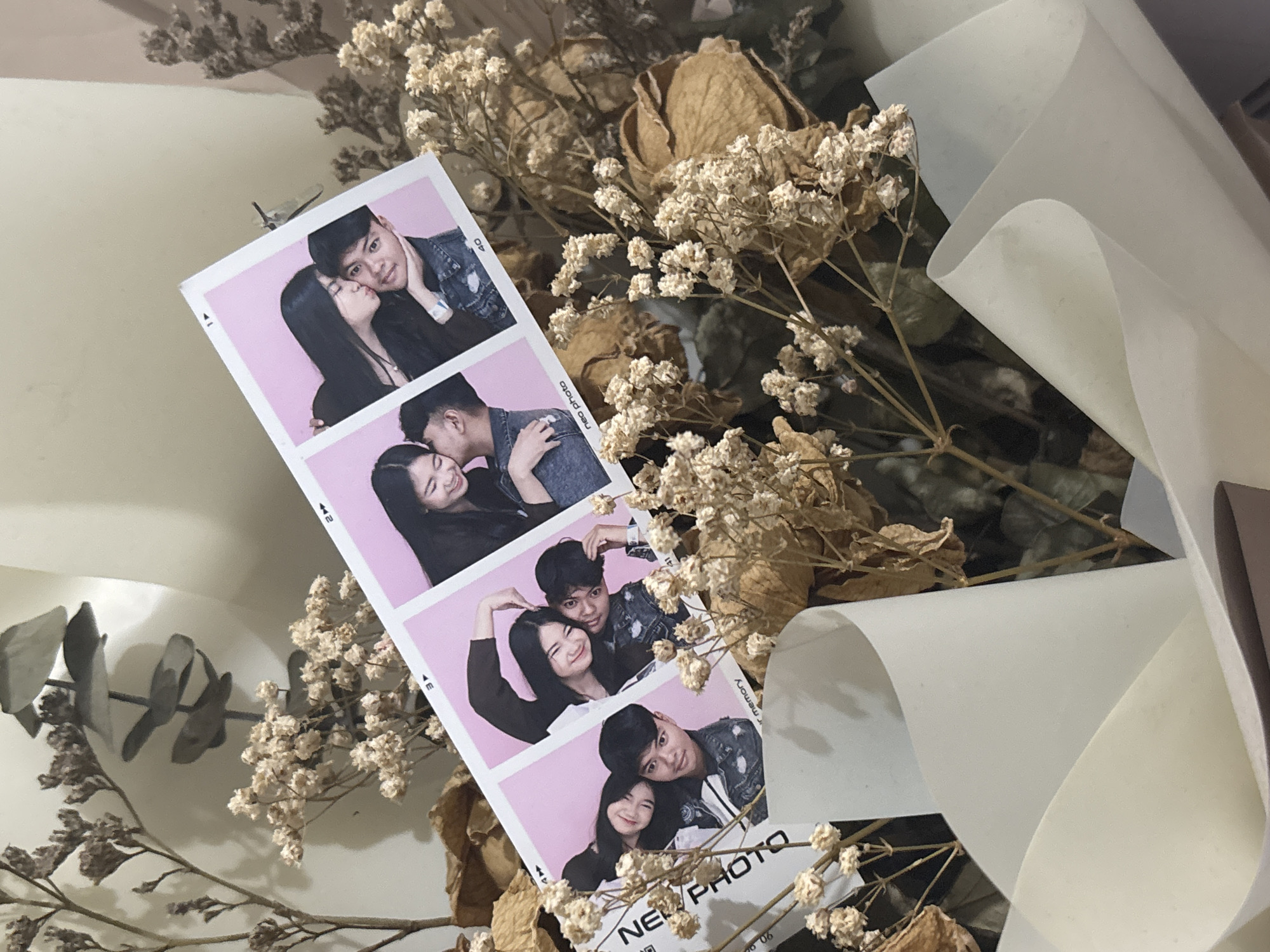 A photo strip of four images of a young Asian couple is displayed over a bouquet of dried flowers and white paper. In the first image, a woman kisses a man on the cheek. In the second image, the couple hugs, and the woman forms a heart shape with her hands. In the third image, the couple smiles closely together. In the fourth image, the woman kisses the man on the cheek. The photo strip has the words "NEO PHOTO" and "your memory" printed on it. The bouquet is composed of light-colored dried flowers and foliage, and is wrapped in white paper. The background is a pale, neutral color, suggesting an indoor setting.