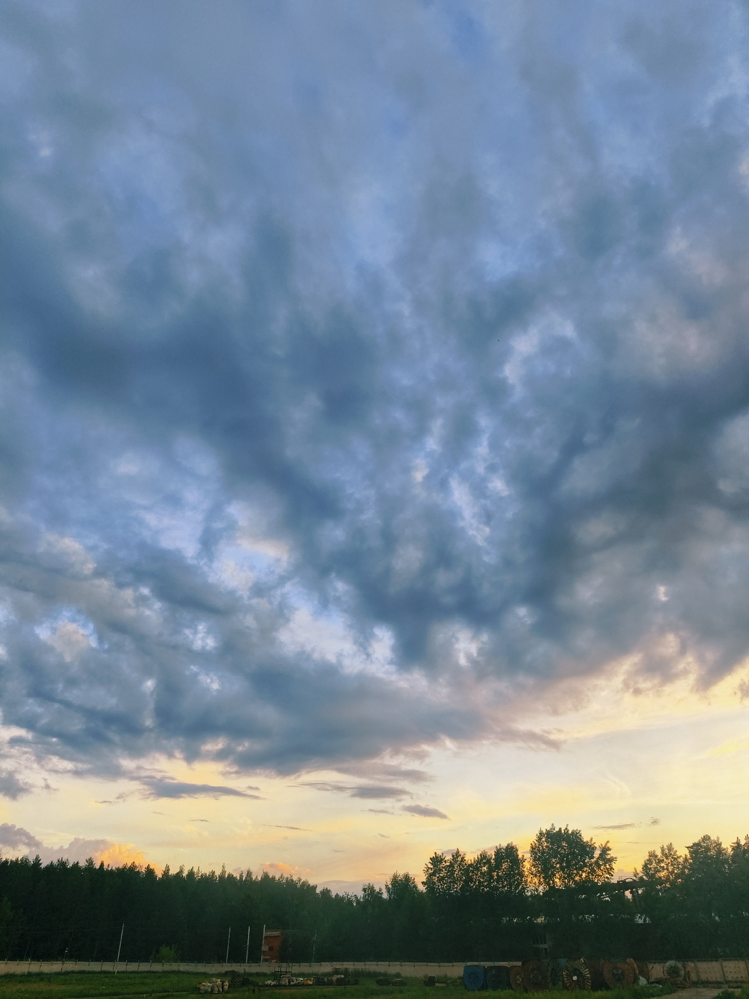The image shows a landscape in Noviy, Russia, during what appears to be late evening or early morning, based on the sky's color. Dominating the upper two-thirds of the frame is a sky filled with dark, heavy clouds transitioning to lighter hues, likely indicating the presence of sunlight behind them. The lower third of the image features a row of silhouetted trees bordering a grassy field. In the field, there are several unidentified objects and a small building. The overall impression is that of a rural or suburban area in Noviy, Russia, at either sunset or sunrise. There are no people visible in the image. The interactions are limited to the natural elements of sky, clouds, trees, and grass.