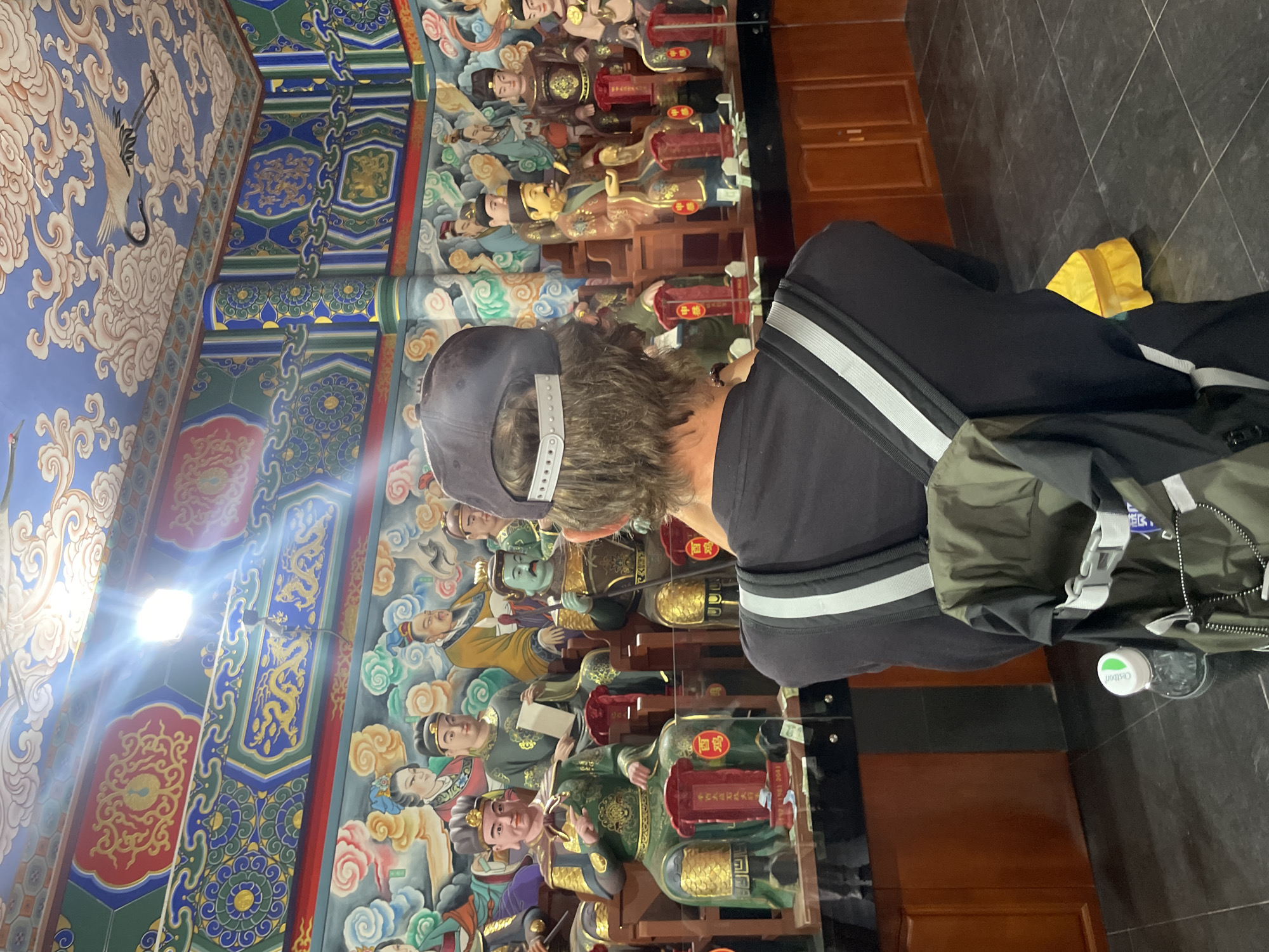 A person wearing a grey baseball cap and a black sweatshirt stands inside a building, possibly a temple or cultural site, in Lintong District, China. The walls are decorated with colorful murals, including images of figures in traditional Chinese clothing. The walls could be decorated with statues. The floor is dark and tiled. The person is wearing a backpack.