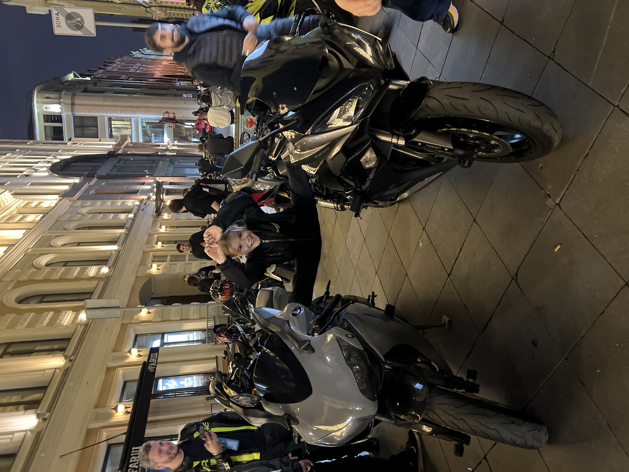 A group of people are gathered on a paved city street at night, possibly for a motorcycle event, in Moscow, Russia. Two prominent sport motorcycles, one gray and one black, are parked side-by-side in the foreground, partially obscuring the pavement. A young person, dressed in black activewear, is performing a seated split between the two motorcycles, with their arms raised and hands near their head, smiling and looking at the camera. Several other individuals are visible in the background, some standing and others seated on a low wall or barrier. They are dressed in casual, warm clothing, suggesting a cool evening. The street is lined with buildings featuring architectural details and illuminated windows, indicative of urban architecture. Overhead streetlights and building lights cast a warm glow on the scene. A sign with Cyrillic lettering, possibly indicating a street name or establishment, is visible on a building to the left.
