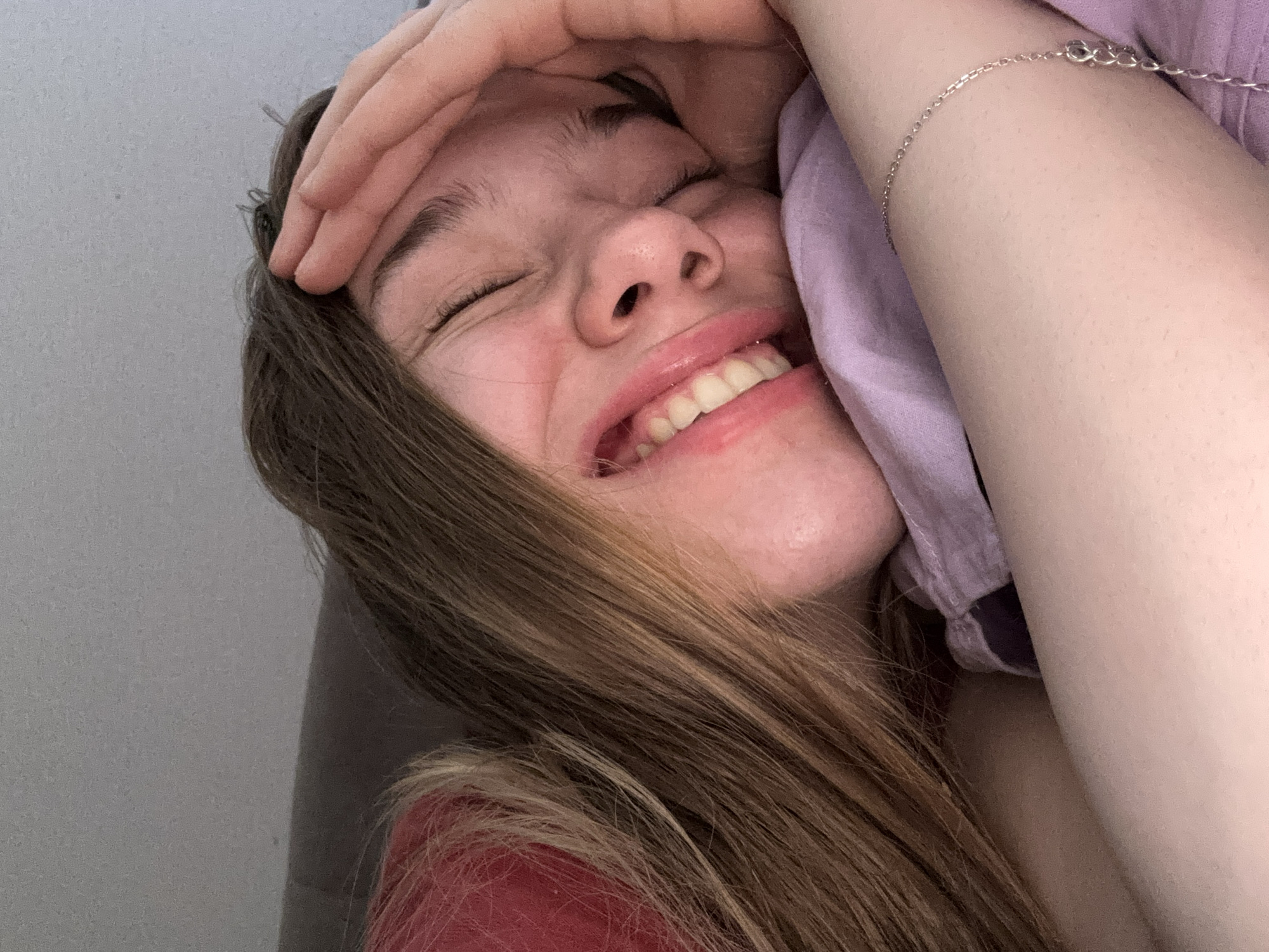 A close-up, overhead, medium shot features a young Caucasian woman with long, wavy, light brown hair. Her eyes are closed, and she has a wide, happy smile, her lips glossed pink. Her right hand is raised, resting on her forehead, partially obscuring her face. She is lying down on her side, her head resting on a purple pillow. A hint of a pink shirt is visible on her left shoulder, and a delicate silver chain bracelet is worn on her right arm. The background consists of a textured grey surface, suggesting an indoor setting, possibly a bed or sofa, with a darker, possibly upholstered, surface behind her head. There is no information available in the image to determine the location in Vsevolozhsk, Russia.