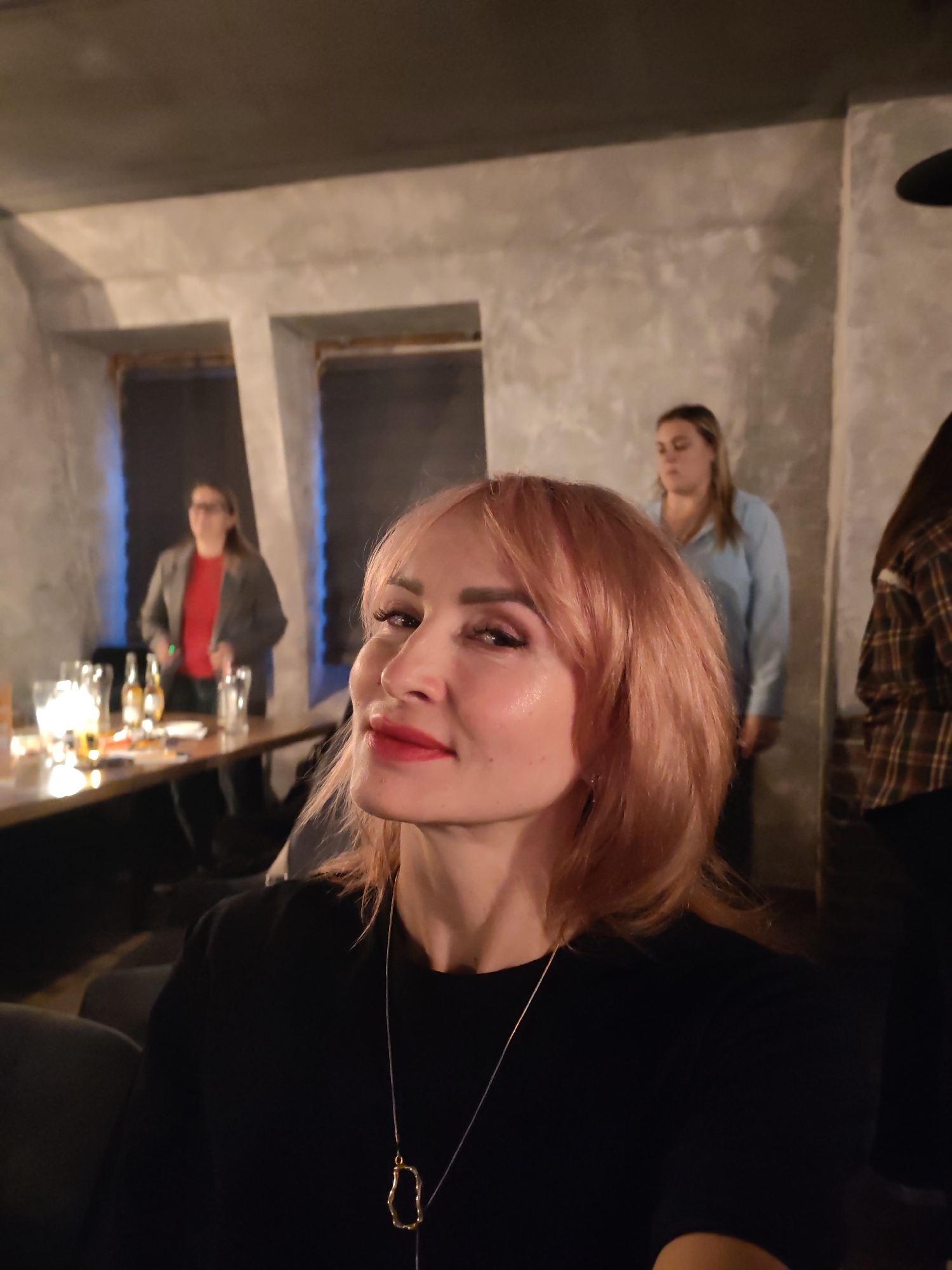 A close-up, eye-level medium shot captures a woman with short, light pink hair styled in soft waves, her head slightly tilted and turned towards the viewer. She wears a black top and a silver necklace with a gold pendant. Her makeup includes bold red lipstick and eye shadow. In the background, a woman in a light blue collared shirt stands with her head down, and another person in a plaid shirt is partially visible on the far right. To the left, another woman in a gray jacket and red shirt is seen behind a table laden with glasses and bottles. The setting appears to be an indoor venue with textured gray walls and muted lighting.