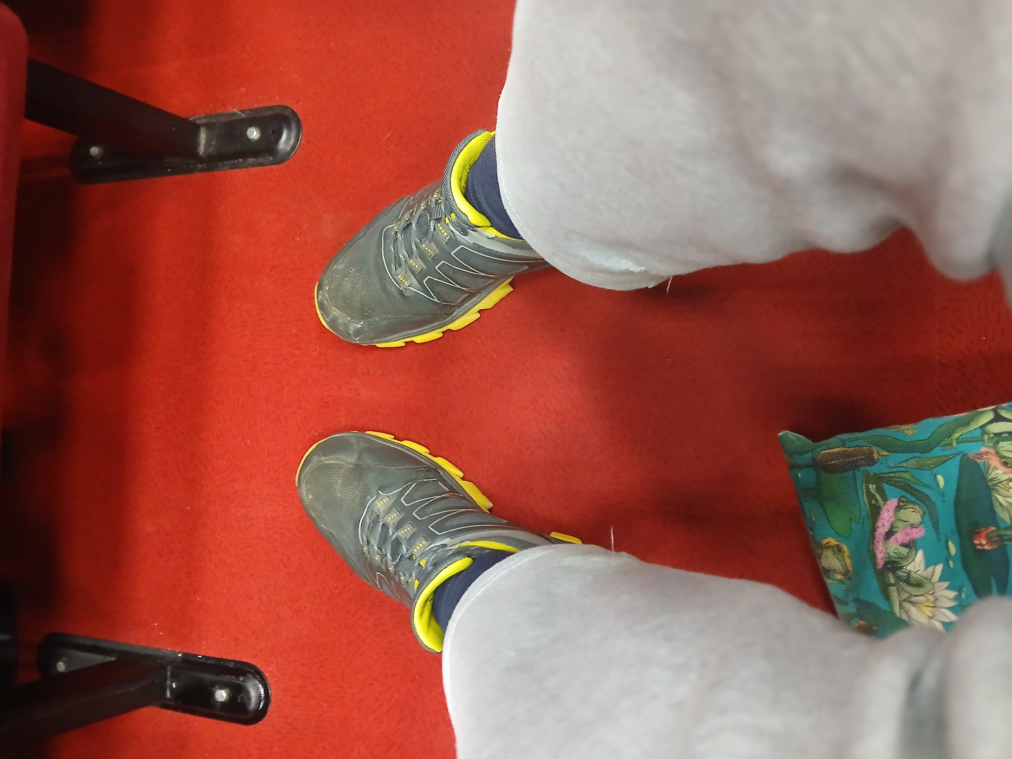 A first-person, high-angle shot reveals a pair of legs clad in grey sweatpants, standing on a vibrant red carpet. The feet are shod in grey and yellow athletic shoes, suggesting an indoor environment. A portion of a black chair leg is visible on the left, while a patterned cushion is situated on the right. The location, Прикамский, Russia, is provided as context.