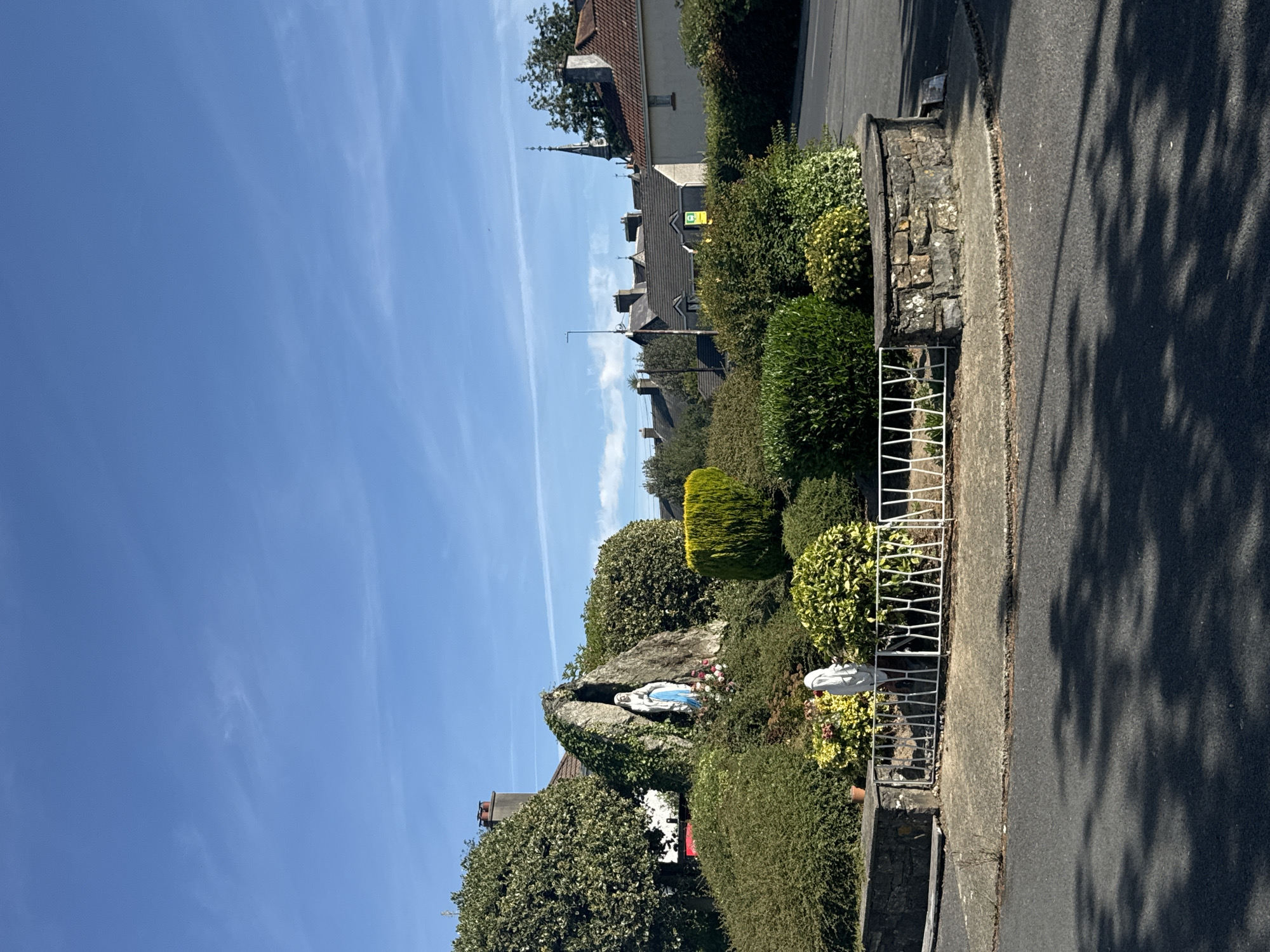 The image shows a religious shrine featuring a statue of the Virgin Mary within a rocky grotto, surrounded by lush greenery and bushes. Another white statue of a figure, possibly also religious, stands near a white metal fence that encloses a section of the shrine. The shrine is situated on a roundabout or traffic island, bordered by a stone wall. In the background, houses with red-tiled roofs and chimneys are visible, along with a church steeple, suggesting an urban or suburban setting. The sky is clear and blue with some wispy clouds, and shadows from trees are cast on the asphalt road in the foreground. This shrine is a well-known landmark in the city of Waterford, Ireland, specifically located on the corner of Ballybeg and Tycor.