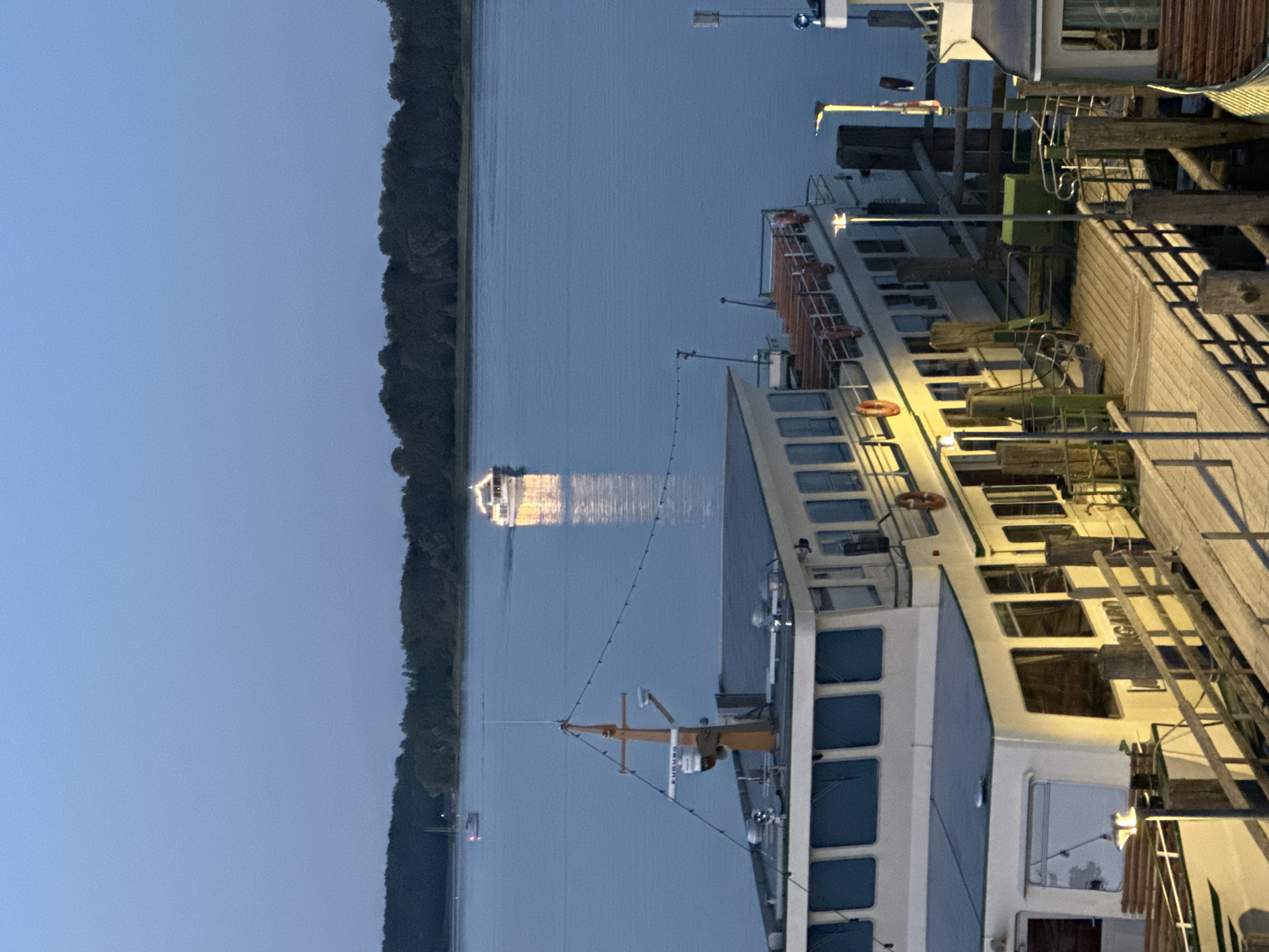 The image depicts a twilight scene at the harbor of Prien am Chiemsee, Germany. A large passenger boat is docked at a wooden pier in the foreground. The boat is illuminated by interior lights and external spotlights, casting a warm glow on the water and the wooden planks of the pier. The boat has multiple decks and numerous windows. A smaller boat is visible further out on the lake, and another larger boat with lights is moving away from the viewer. The background is dominated by a dark, tree-lined shore. The sky is a clear, deep blue, indicative of dusk. The scene is tranquil and serene, with the gentle lapping of water and the distant hum of engines being the only suggested sounds.