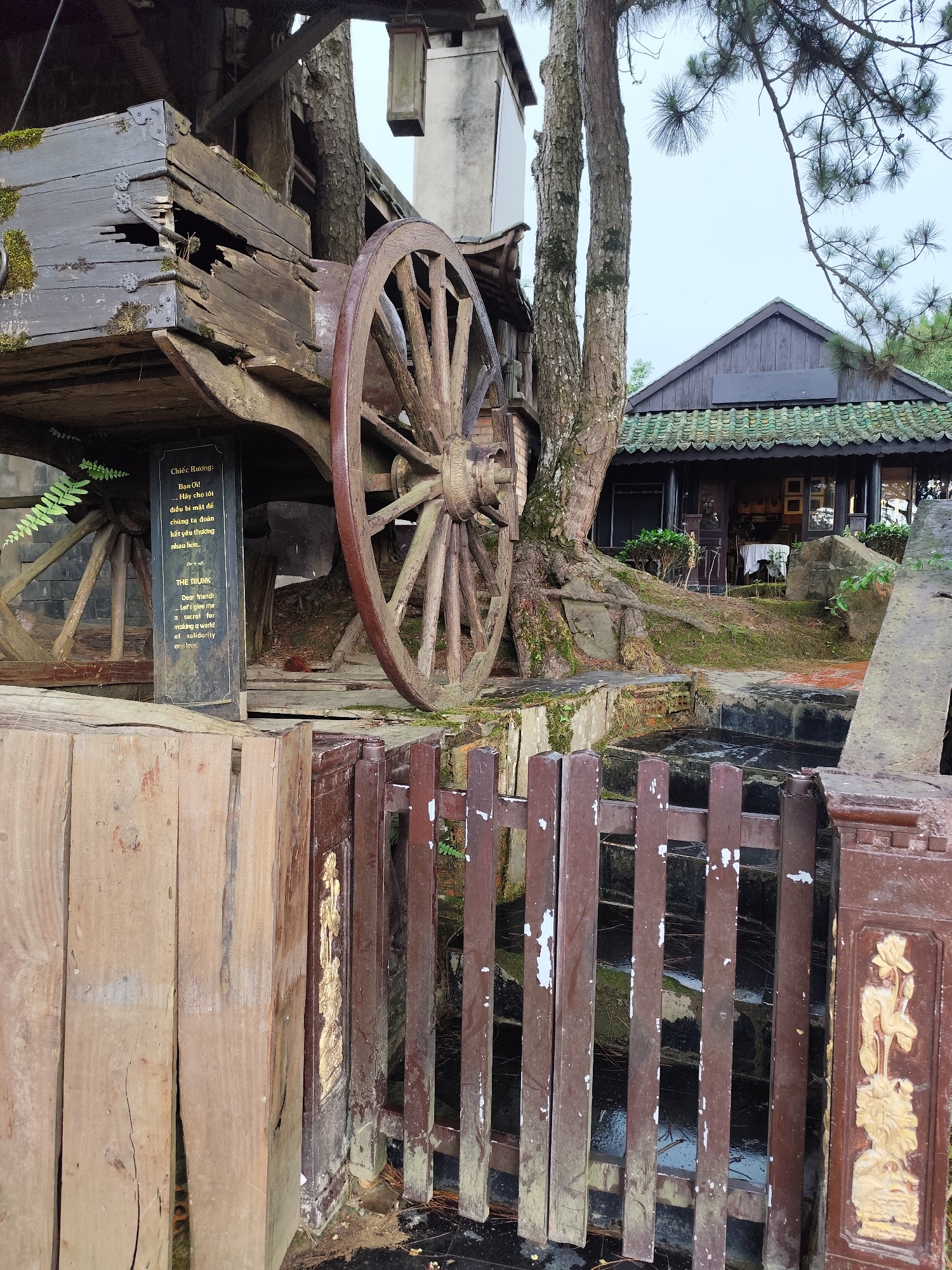 The image shows an old wooden cart with large wheels, positioned in front of a building with a green-tiled roof, typical of architecture in Lam Vien - Da Lat, Vietnam. A sign is affixed to the cart, featuring text in Vietnamese and English, titled "The Trunk" and "Chiếc Rương", respectively. The sign mentions a secret for making a world of solidarity and love. A decorative fence sits in the foreground, with carved designs. A tree grows close to the cart and the building in the background is partially obscured by trees and the cart itself.