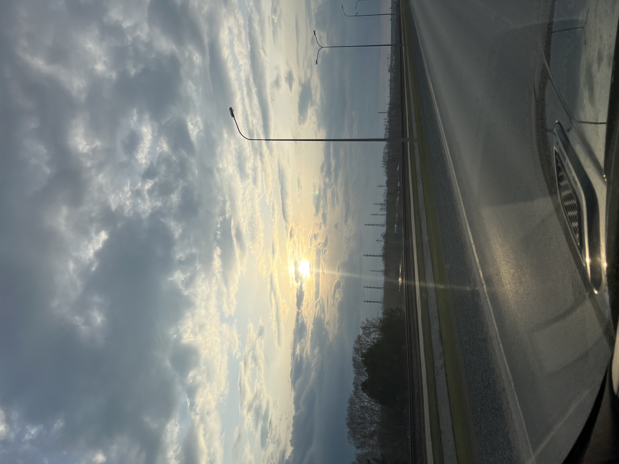 The image captures a dynamic outdoor scene from the perspective of a vehicle, looking towards the horizon during either sunrise or sunset. The sky dominates the upper and left portions of the frame (when correctly oriented), filled with a mix of fluffy white and grey clouds, some illuminated brightly by the sun and others in shadow. The sun itself is a brilliant, low-lying orb near the center, casting a strong glare and warm light through a break in the cloud cover.
Below the sun and along the horizon, a dark line of silhouetted trees is visible, followed by a series of power line towers stretching into the distance. To the right, an asphalt road with visible white lane markings extends horizontally. Several tall streetlights with distinctive curved arms line the road, their forms silhouetted against the brighter sky. On the far right, the dark, reflective surface of a car's hood and front fender are visible, indicating the vantage point from inside a moving or parked vehicle.
No people or explicit text are visible in the scene. The low sun position and warm light suggest either early morning or late afternoon, while the scattered clouds create an atmospheric sky. The setting appears to be a road or highway, likely near a semi-urban or rural area in Chaikovsky, Russia, given the industrial structures and natural elements.