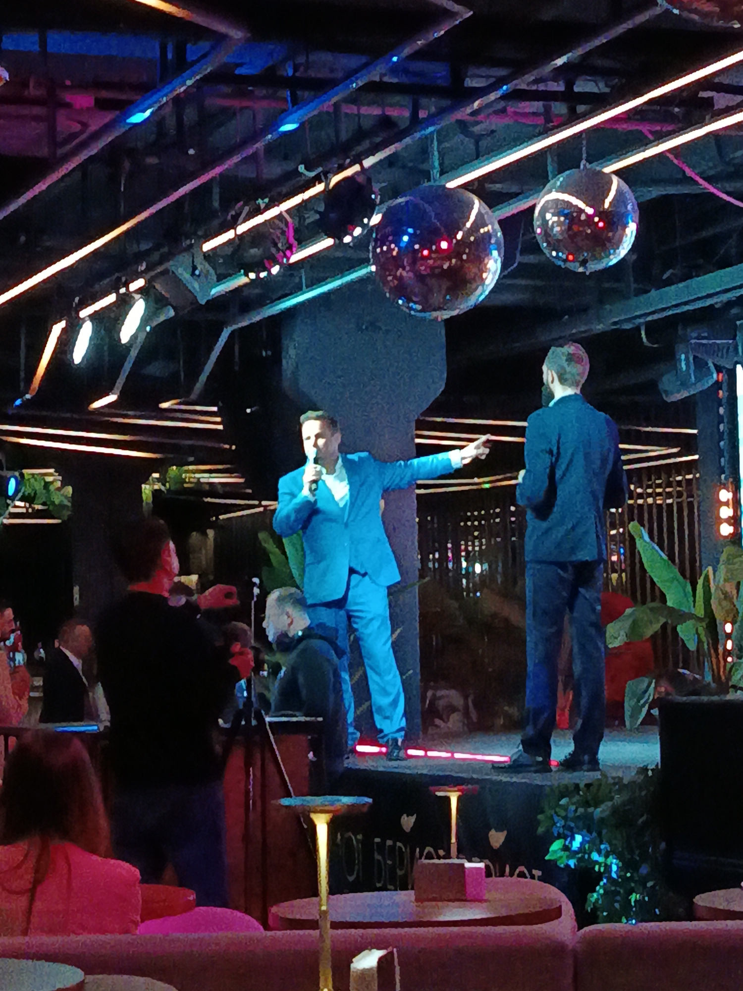 The image shows a man in a blue suit on a stage, speaking into a microphone and gesturing with his right hand. He is standing next to another man in a dark suit, who is facing away from the camera. The stage is decorated with plants and lighting, and there are disco balls hanging from the ceiling. The audience is seated at tables with pink tablecloths. The location is likely a nightclub or event space in Moscow, Russia.