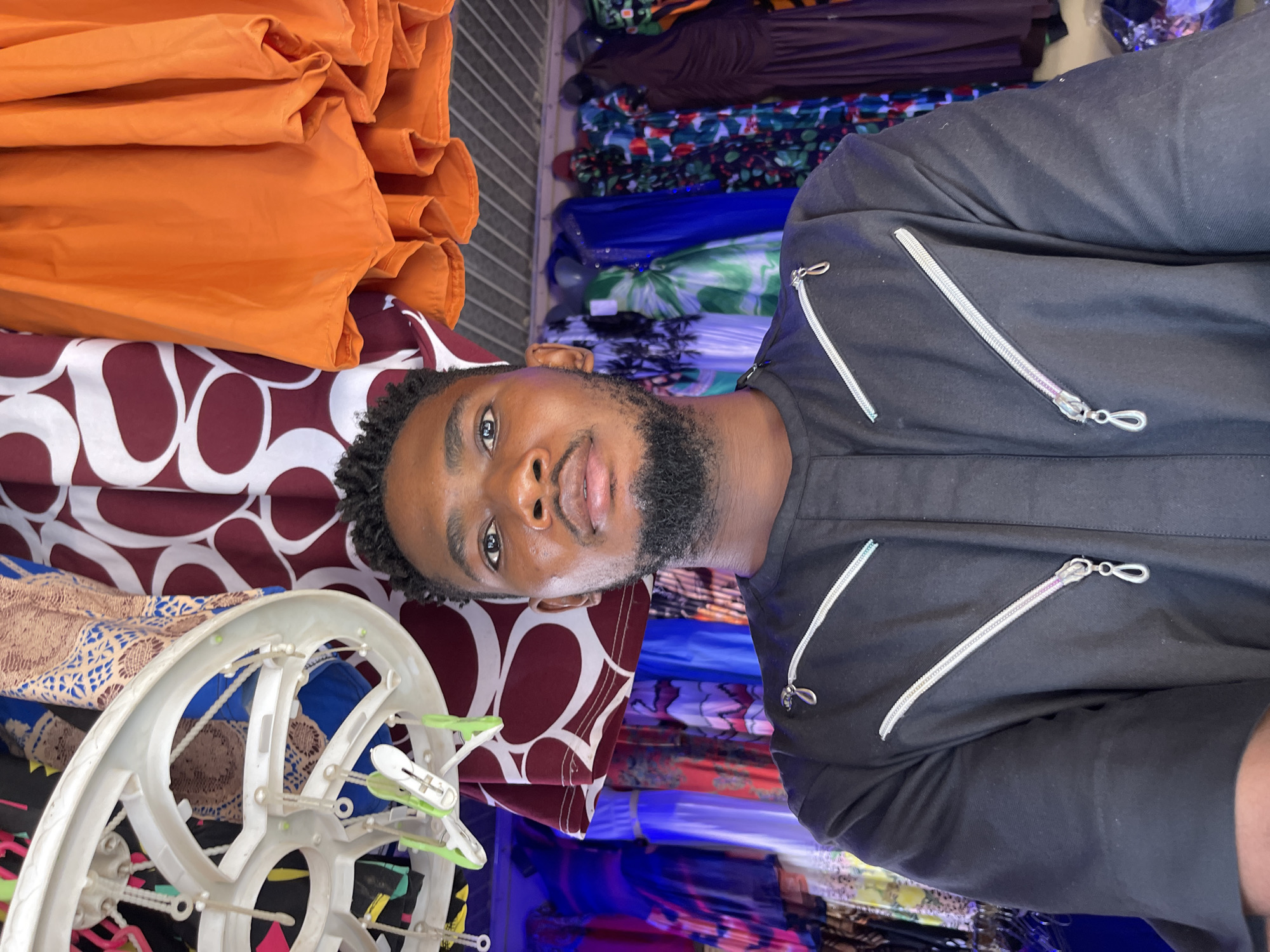 The image shows a man in a black shirt with decorative silver zippers, positioned in what appears to be a retail setting in Samaru, Nigeria. The backdrop consists of textiles and fabrics of varying colors and patterns. There is an orange fabric, a fabric with white and maroon oval shapes, and a rack with folded fabric in the background. To the left, there is a white circular clothing rack with green clothespins. The man faces the camera.
