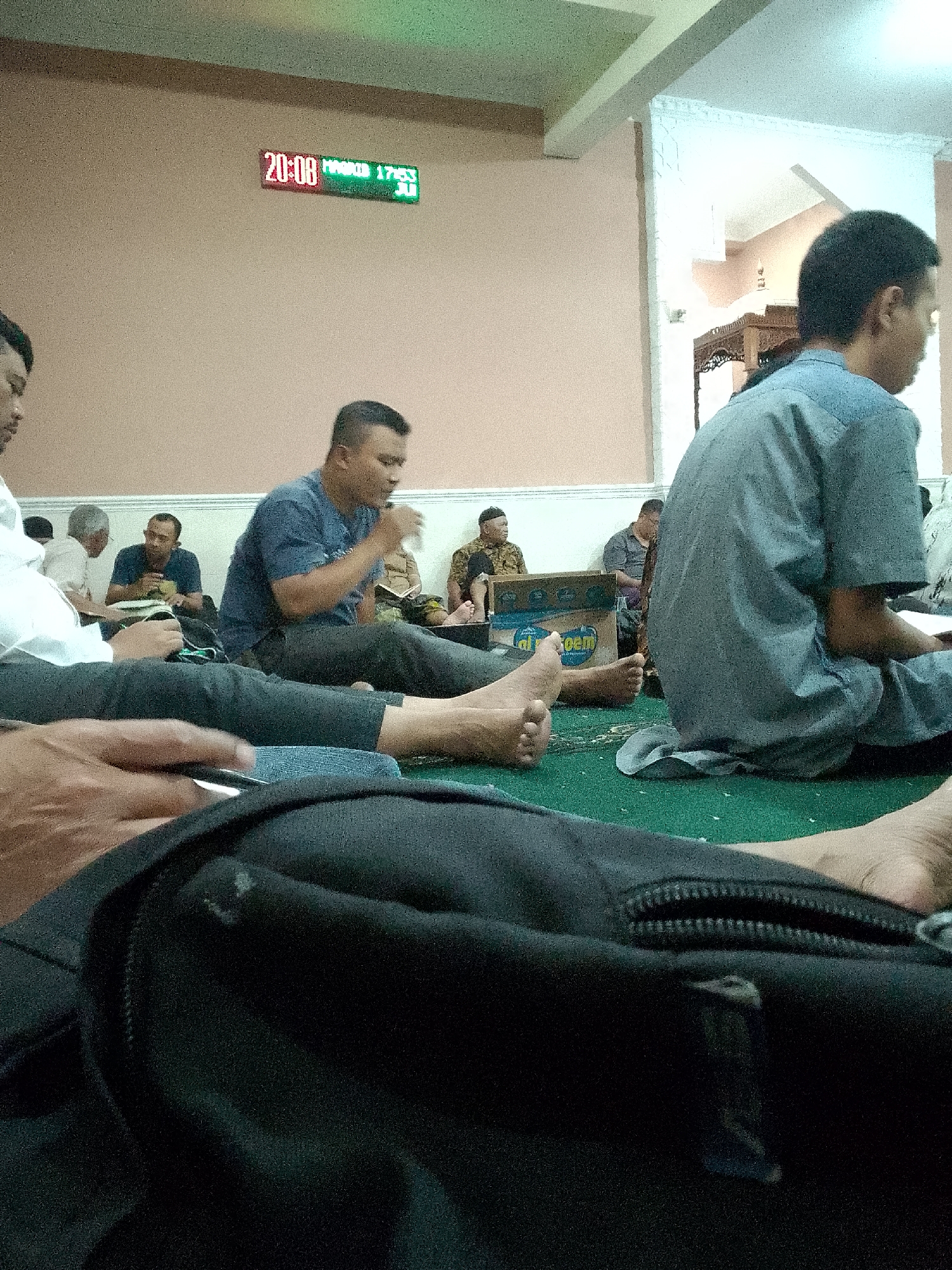 The image captures an indoor scene, likely a mosque or prayer hall, based on the presence of multiple individuals sitting on the floor and the religious paraphernalia visible. In the background, a digital clock displays the time as "20:08" and possibly a date or prayer time, "MAGRIB 17:53 JUL". The room features a light-colored ceiling and walls, with a large white pillar visible on the right side. A green carpet covers the floor where several men are seated. In the foreground, a black backpack is prominently displayed, partially obscuring the lower portion of the image. Several men are depicted in various states of sitting and interacting. One man in the foreground, wearing a blue t-shirt and dark pants, is leaning forward and holding a drink. Another man, further back and to the left, is wearing a white shirt and appears to be reading. Other individuals are also seated on the carpet, some engaged in conversation or prayer. The overall atmosphere suggests a gathering of people for religious purposes. The image was taken in Kertajaya, Indonesia.