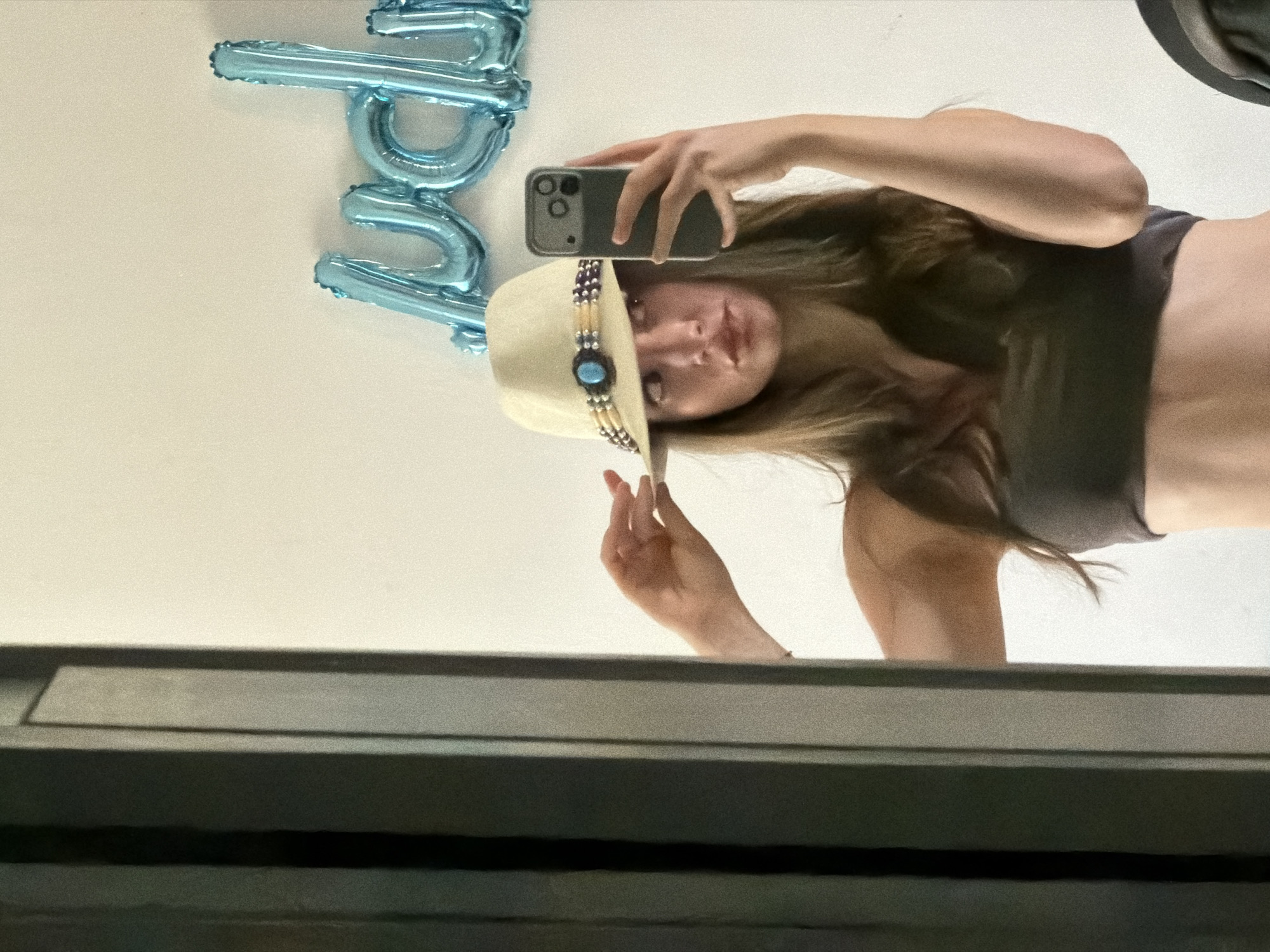 A young woman is taking a selfie in a mirror, her head tilted to the side. She is wearing a straw cowboy hat adorned with blue and silver beads and a large blue stone. Her hair is long and brown, falling over her shoulders and obscuring part of her face. She is also wearing a dark grey sports bra. In the background, blue foil balloons spelling out "Happy B." are visible. The reflection shows her torso and arm, and a portion of what appears to be a window frame or ledge in the foreground. The lighting suggests an indoor setting, potentially during daylight hours. No explicit weather or time of day cues are present, but the overall impression is casual and possibly celebratory. The context provided suggests the location is Sharm El Sheikh, Egypt.