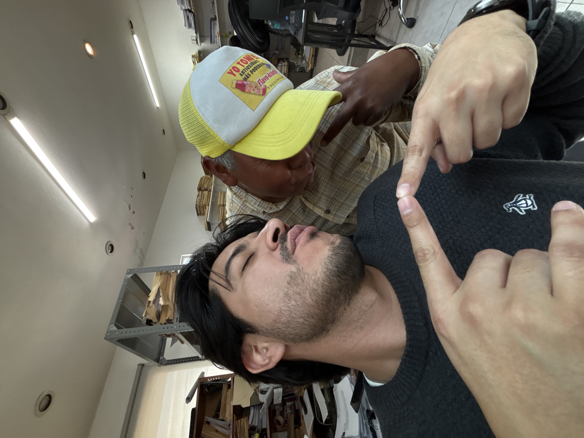 Two men are in an office setting, in what appears to be the city of Puebla, Mexico. The man on the right is wearing a gray sweater with a penguin logo and a watch. His right hand is pointing at himself. The other man is wearing a yellow and white hat that reads “Yo Tomo antioxidantes más poderosos” and a plaid shirt. He is pointing at the man with the sweater’s head. There are shelves with paper and objects visible in the background.