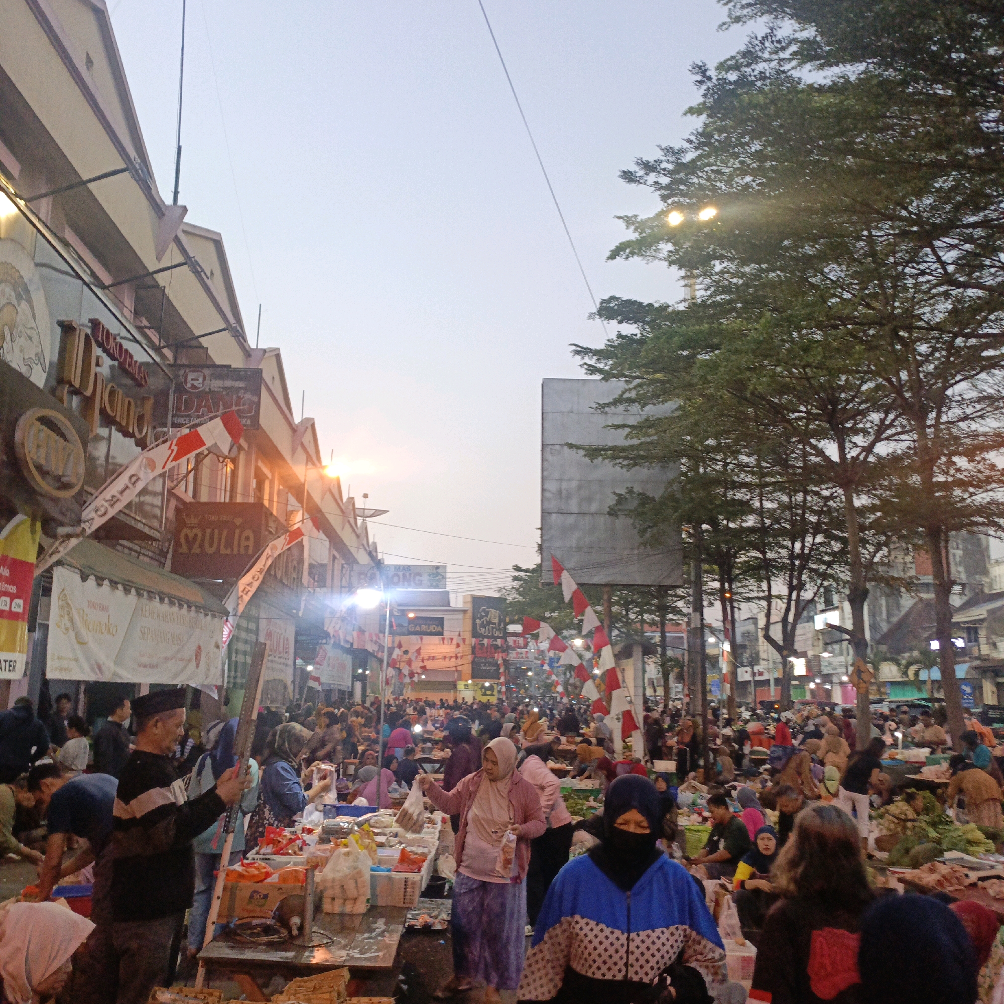 The image depicts a busy outdoor market scene in Salatiga, Indonesia, likely during the evening or early morning given the lighting. The foreground is dominated by market stalls filled with various goods, including what appears to be food items, packaged goods, and possibly textiles. Several people are visible engaged in shopping and selling.
In the midground, a dense crowd of people navigates through the narrow aisles between the stalls. Many are wearing traditional Indonesian attire, including headscarves (hijab) for women and peci caps for men. The architecture of the buildings lining the street is visible, featuring shopfronts with signage, some of which are illuminated. Red and white Indonesian flags are displayed prominently, suggesting a national celebration or event.
The background shows more buildings and trees, with the sky being a pale, overcast hue. The overall impression is one of a vibrant and bustling community gathering for commerce. The presence of numerous individuals, diverse goods, and the lively atmosphere are key characteristics of this Indonesian market.