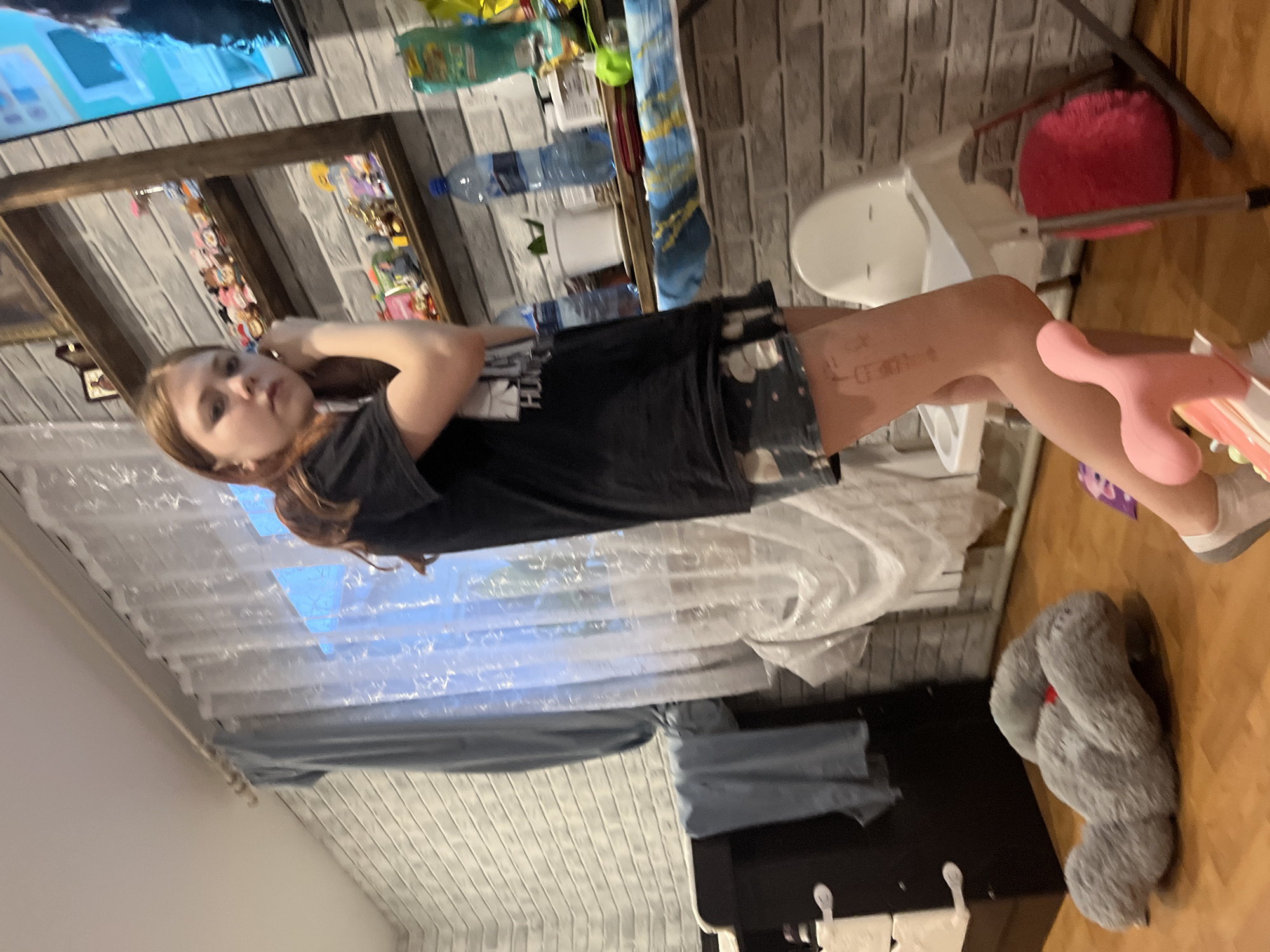A girl in a black shirt and patterned shorts lies on her side in a room. A window with a white curtain is visible behind her. A teddy bear rests on a wood floor next to a black dresser with white knobs. A high chair with a pink seat cushion sits against a brick wall. Bottles and other items are visible on a shelf. The room's specific location within Vesyoloye, Russia, cannot be determined from the image.