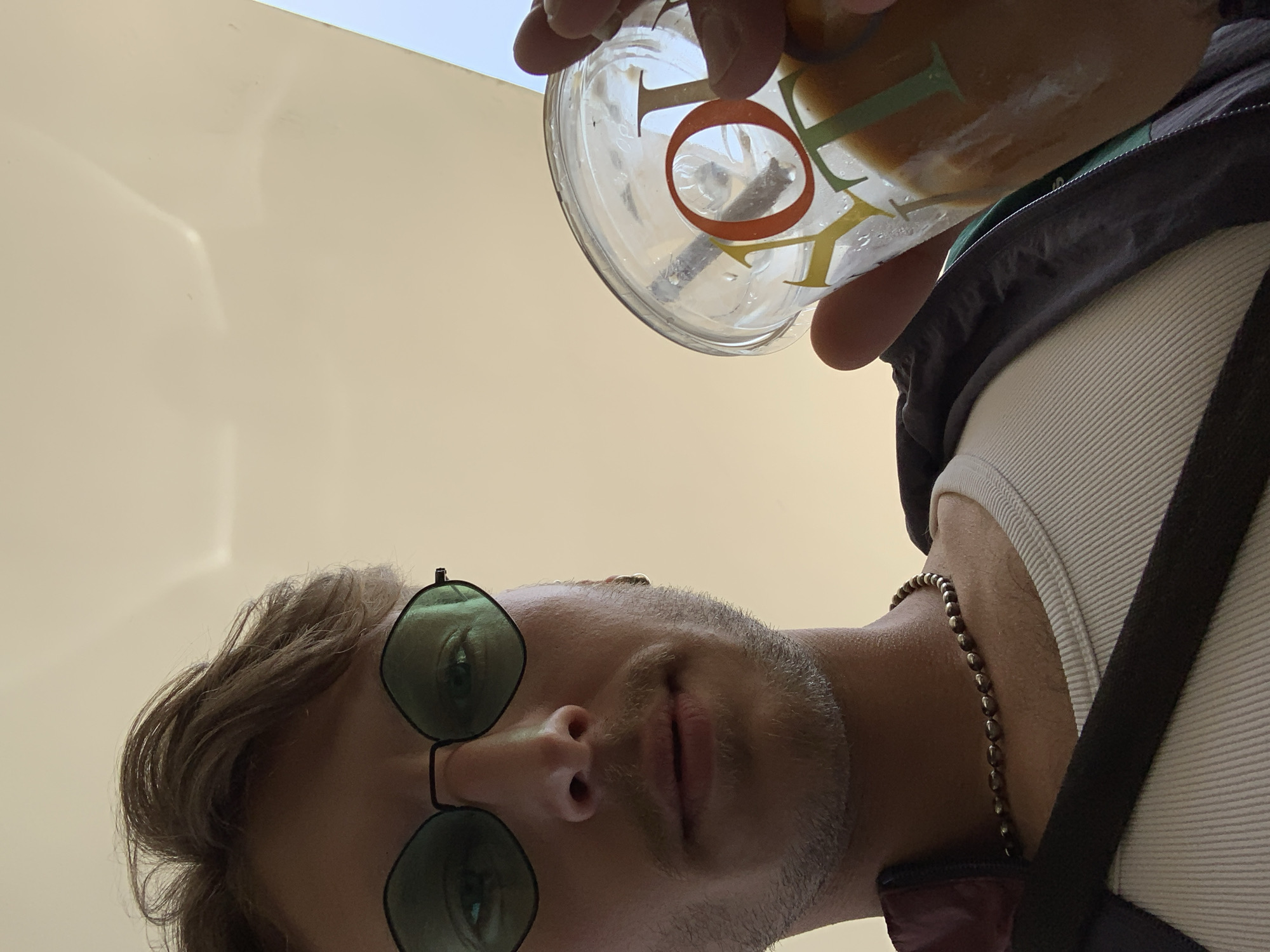A low-angle, upward-facing shot captures a person, likely male, holding a beverage. The subject has messy brown hair and wears round, green-tinted sunglasses. A light stubble or beard covers their face, and a neutral expression is discernible. They are dressed in a white ribbed tank top, over which a dark jacket or hoodie is visible. A beaded, metallic-looking necklace is worn around their neck.
The person holds a clear plastic cup containing a light brown, milky beverage with a straw. On the cup, the letters "TO" are clearly visible in colorful fonts, with the "O" featuring a prominent orange outline. Other partial letters, possibly forming a brand or word, are also present. The background is predominantly a plain, light beige surface, likely a wall or ceiling, with a small sliver of clear blue sky visible in the upper right corner. The lighting is bright, creating subtle reflections on the beige background. The perspective suggests the camera is positioned low, looking up at the subject. This image is set in Tel-Aviv, Israel.