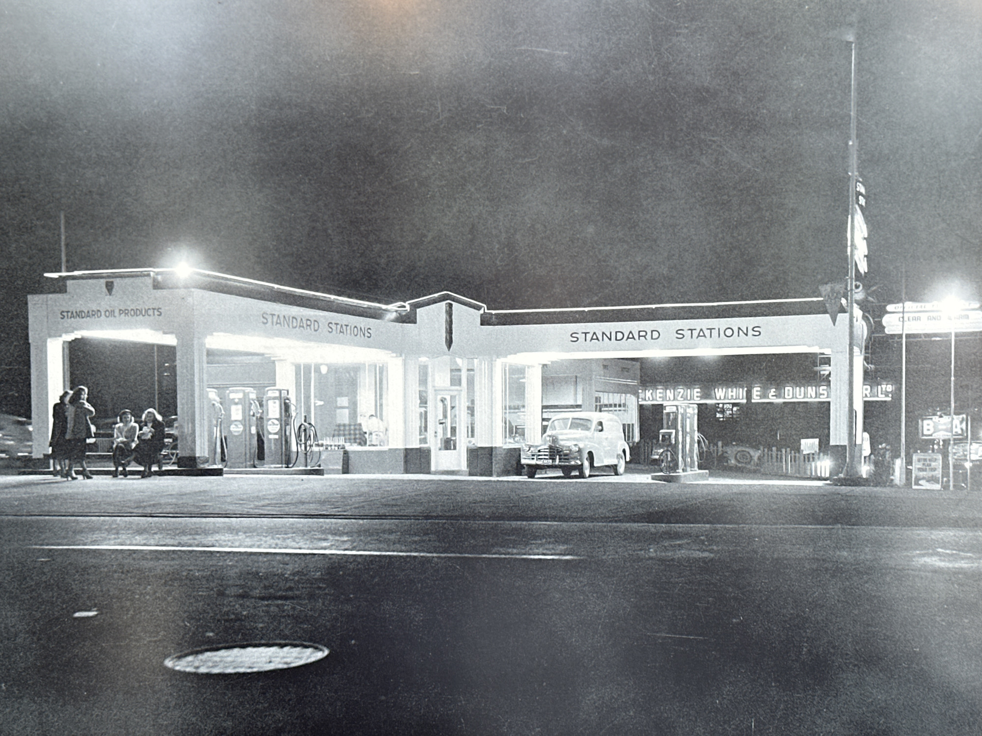 The photograph captures a Standard Oil station in Langley, Canada, likely around the mid-20th century based on the vehicle and clothing styles. The station features a multi-section canopy with "STANDARD OIL PRODUCTS" and "STANDARD STATIONS" prominently displayed. Several gas pumps are visible under the canopy. A vintage panel truck is parked near the right side of the station. Behind the station building, additional signage reads "KENZIE WHITE & DUNS RLTO". The surrounding area includes signs for other businesses, including one for "BA" (British American Oil). On the left side of the station, a group of four women are sitting on a bench. The ground is paved and has a manhole cover visible. The scene is illuminated by lights from the station and nearby signs, creating a contrast with the dark sky.