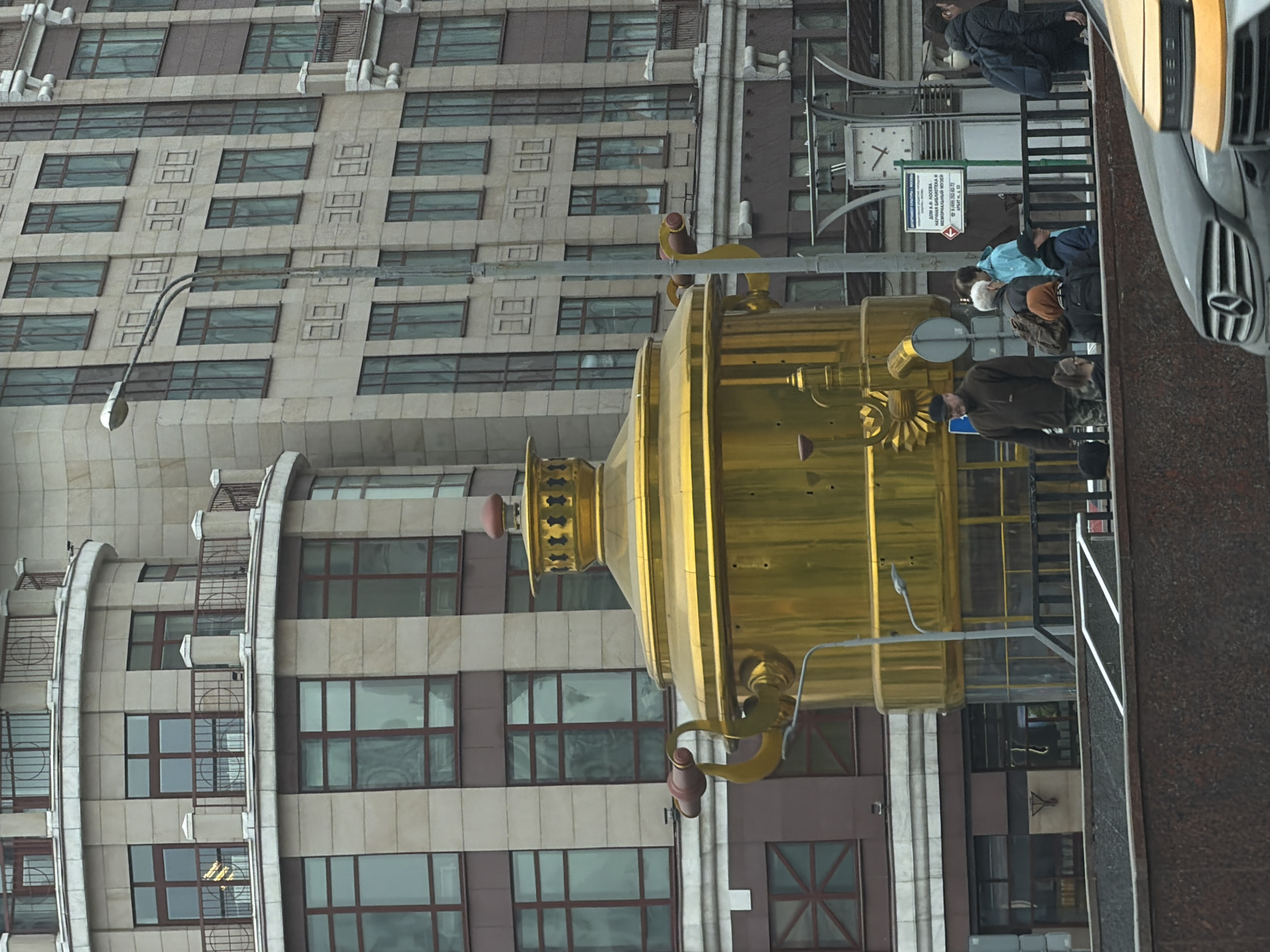 The image depicts an urban street scene in Moscow, Russia, specifically on Arbat Street near the Losev House (Dom Loseva) at address 33. The central subject is a large, ornate, golden samovar-shaped monument situated in the mid-ground, reflecting the overcast sky. Behind the samovar, a multi-story building with a rounded corner and numerous windows framed in dark material is visible on the left, featuring balconies with dark metal railings. To the right and extending into the background, another tall, light-colored building with repetitive square decorative elements and dark-framed windows rises.
In the mid-ground, a group of approximately seven to eight pedestrians is present behind a dark metal railing and a granite-like wall. Individuals are dressed in jackets and long sleeves, suggesting cooler weather. Notable figures include a man in a dark jacket and cap carrying a backpack, an older woman with grey hair and a backpack, and a person in a light blue jacket. Other individuals are partially obscured. A green and white sign, partially legible, indicates "ДОМ ЛОСЕВА НАУЧНАЯ БИБЛИОТЕКА..." (Losev House Scientific Library...) and "АРБАТ, 33". A large square clock with Roman numerals is visible mounted on a decorative metal structure to the right of the samovar. A grey street lamp with a double arm extends vertically from the mid-ground.
In the immediate foreground, the front section of a dark grey Mercedes-Benz sedan is visible on the left, adjacent to the front portion of a yellow taxi on the right. The sky is uniformly overcast, indicating diffuse daylight conditions.