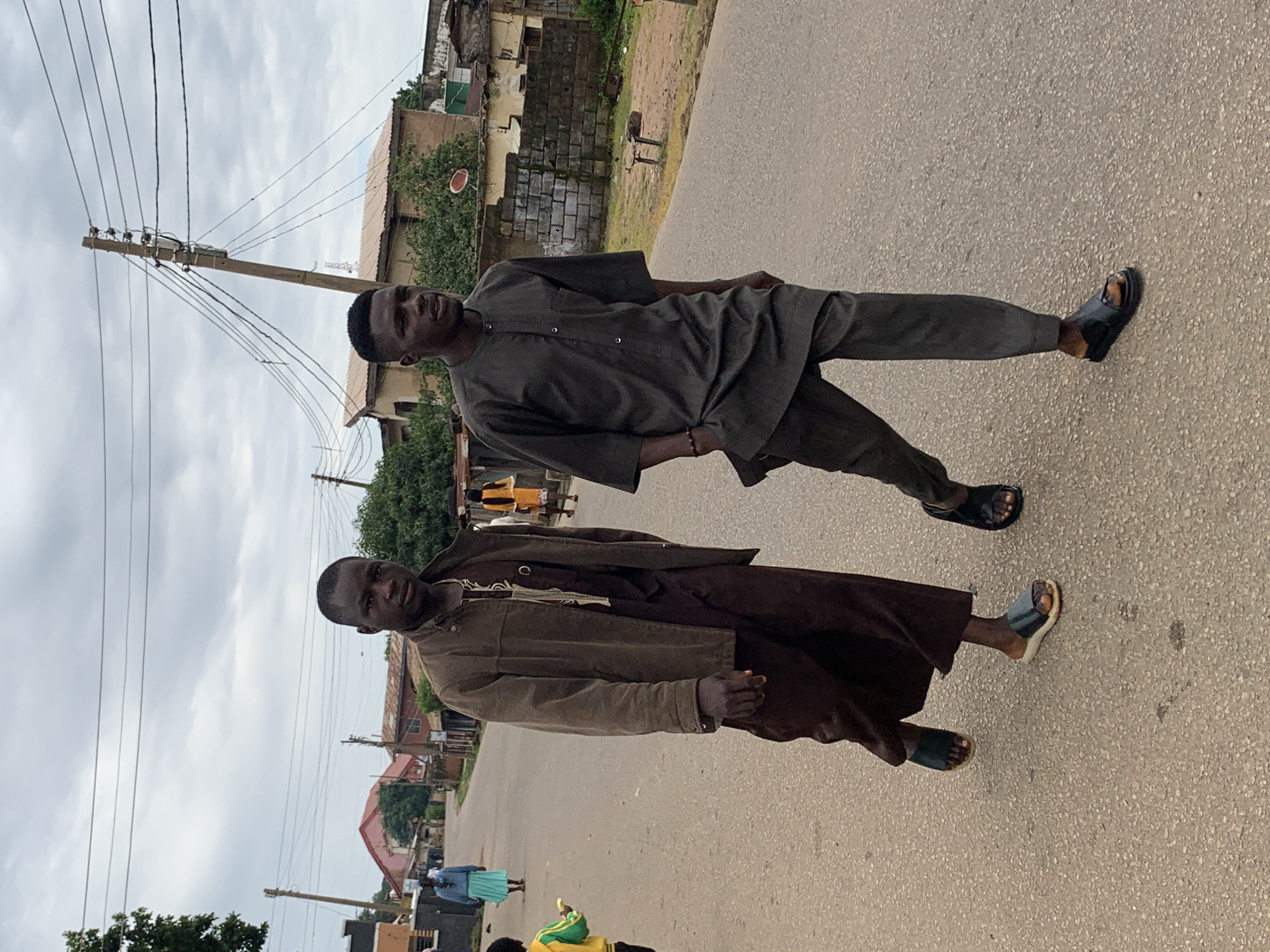 Two young Black men are walking on a paved street in Jos, Nigeria. The man on the left wears a dark brown traditional outfit with a matching long-sleeved tunic and pants. He also has a brown jacket over his tunic and wears dark-colored sandals. The man on the right wears a dark grey, loose-fitting tunic and matching pants, also with dark-colored sandals. They are walking away from the camera and slightly towards the left side of the frame.
In the background, there is a dusty street with a few scattered buildings, some appearing to be residential. There are also utility poles with numerous wires crossing overhead, indicating an urban or semi-urban setting. Other people are visible in the distance, including a woman in a light blue headscarf and a long turquoise skirt, and a child in a yellow and green jacket. The sky is overcast with clouds. The overall impression is that of a casual day on a street in Jos.
