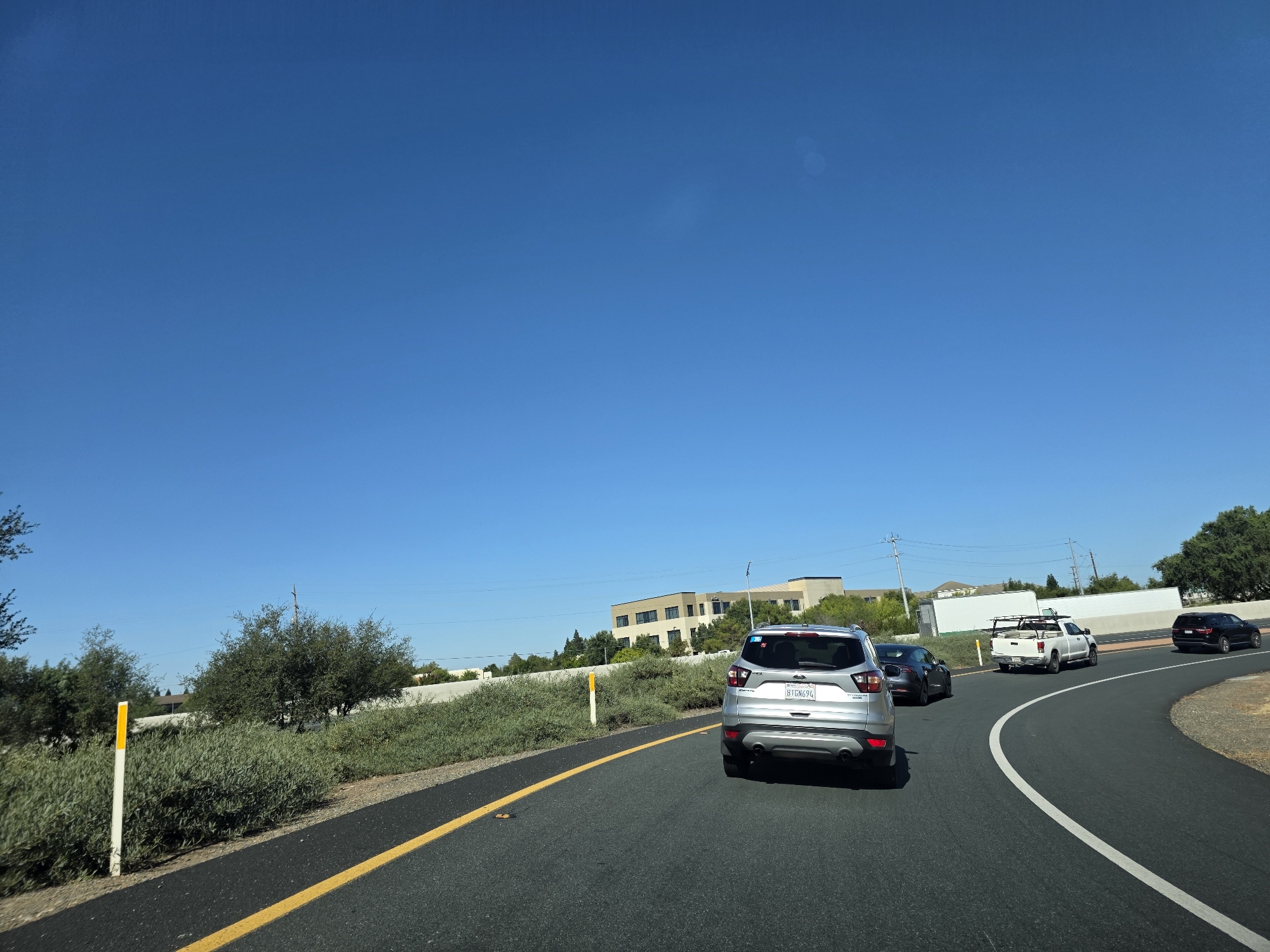 The image depicts a bright, sunny day with a clear blue sky, suggesting it is mid-morning or afternoon. The scene is a highway in Elk Grove, California, with cars traveling along a paved road. The road has a yellow lane marker and a white dividing line. On the left side of the road, there are green bushes and trees, along with a yellow and white reflective marker post. In the distance, there are several buildings, appearing to be offices or a business park.
Several vehicles are visible on the road. Closest to the viewer is a silver SUV, followed by a dark-colored sedan. Further ahead, there is a white pickup truck with a rack on its bed, and another dark SUV. The vehicles appear to be in moderate traffic, moving along the highway.
One of the visible license plates on the silver SUV reads "8TGN694". The overall impression is a typical drive on a sunny day in a suburban or commercial area. There are no apparent signs of urgency or distress; the scene is calm and ordinary. The clear sky and bright sunlight indicate good weather conditions.