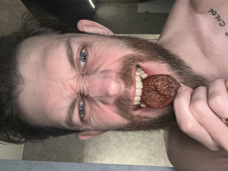 A close-up, front-facing image depicts a Caucasian male with dark, disheveled hair, a full brown beard, and blue eyes. He is shirtless and exhibits an exaggerated facial expression, characterized by furrowed brows, a wrinkled nose, and lips pulled back to expose his upper and lower teeth. He holds a single, shriveled, dark reddish-brown fruit, consistent with a dried apricot or prune, between his front teeth, supported by his left hand. A gold-colored ring is visible on the ring finger of his left hand. A partial tattoo, appearing to show Cyrillic or stylized Latin script (e.g., "CHOL"), is visible on his upper right chest. The background is an interior space with a light, textured wall on the left and a darker, possibly reflective surface with a vertical light element on the right. The specific location within Saint Petersburg, Russia, cannot be determined from the visual information provided.