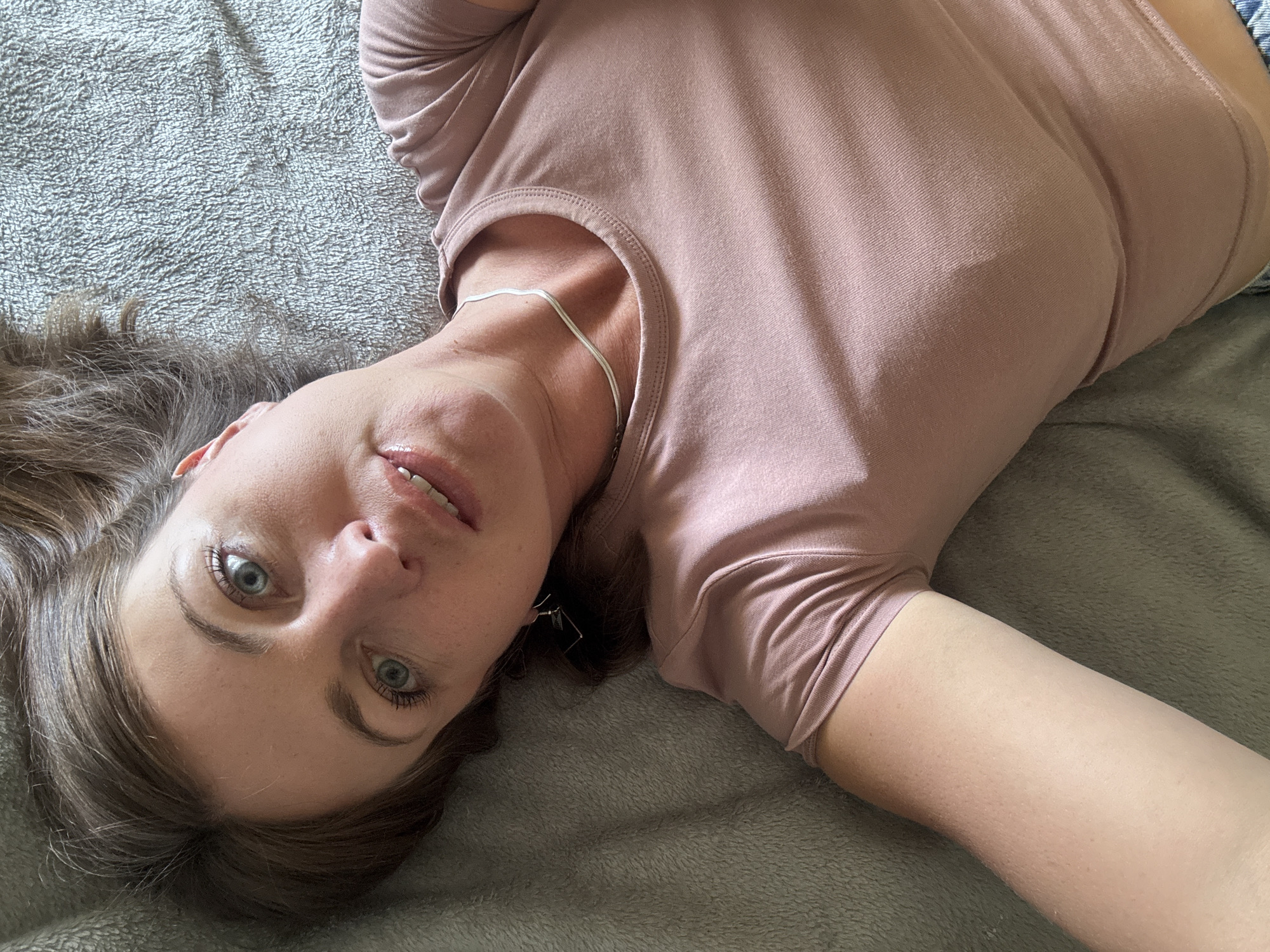 A woman with light brown hair and blue eyes lies on her back on a textured grey surface, wearing a pale pink t-shirt and a silver necklace. She is looking up at the camera with her mouth slightly open. Her hair is spread out around her head. The scene appears to be indoors, possibly in a bedroom or living room. No specific location in Saint-Claude, France can be determined from the image.