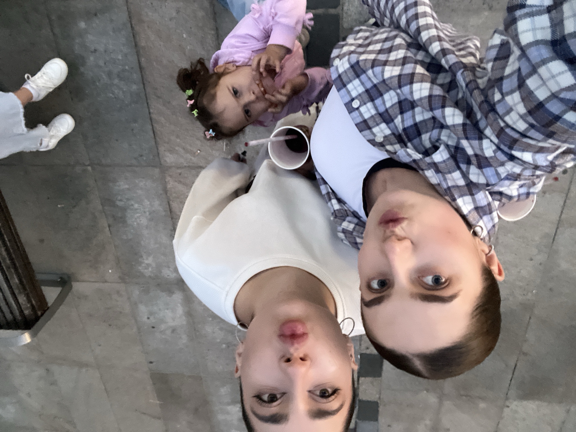 In the city of Lublin, Poland, on an outdoor paved walkway, a group of three individuals are captured in a high-angle selfie. The foreground is dominated by the faces of two young women. The woman on the left, closest to the camera, has her lips pursed. She is wearing a white sweatshirt and a silver hoop earring. To her right and slightly lower, another young woman is visible. She has wide-open blue eyes and is looking directly at the camera. She is wearing a blue and white plaid shirt over a white t-shirt. A white paper cup with a red straw is partially visible near her.
Behind and to the right of the two women, a young girl is also in the frame. She has dark hair styled with colorful clips and is wearing a light purple hooded jacket. She is actively eating something. Her hands are near her mouth, and she is holding pink food.
The background consists of the textured grey pavement, which appears to be made of large stone tiles. In the upper left corner, a portion of a metal bench with wooden slats is visible, suggesting the selfie was taken in a public area, possibly a park or street. The overall lighting suggests it is daytime.