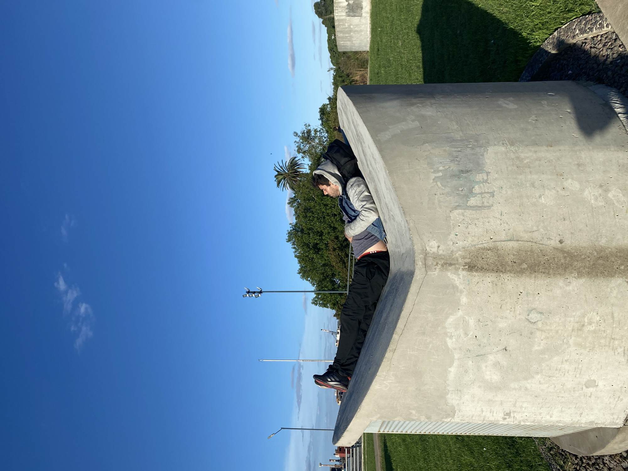 A man is sitting inside a large, concrete, tunnel-like structure in Buenos Aires, Argentina. He wears black pants, a grey hooded jacket, a denim vest, and black-and-red sneakers. He faces left, inside the tunnel, with a blue sky and some trees in the background. The tunnel is part of a series of similar concrete structures located on a grassy area, under a blue sky. The scene appears to be outdoors, in daylight.