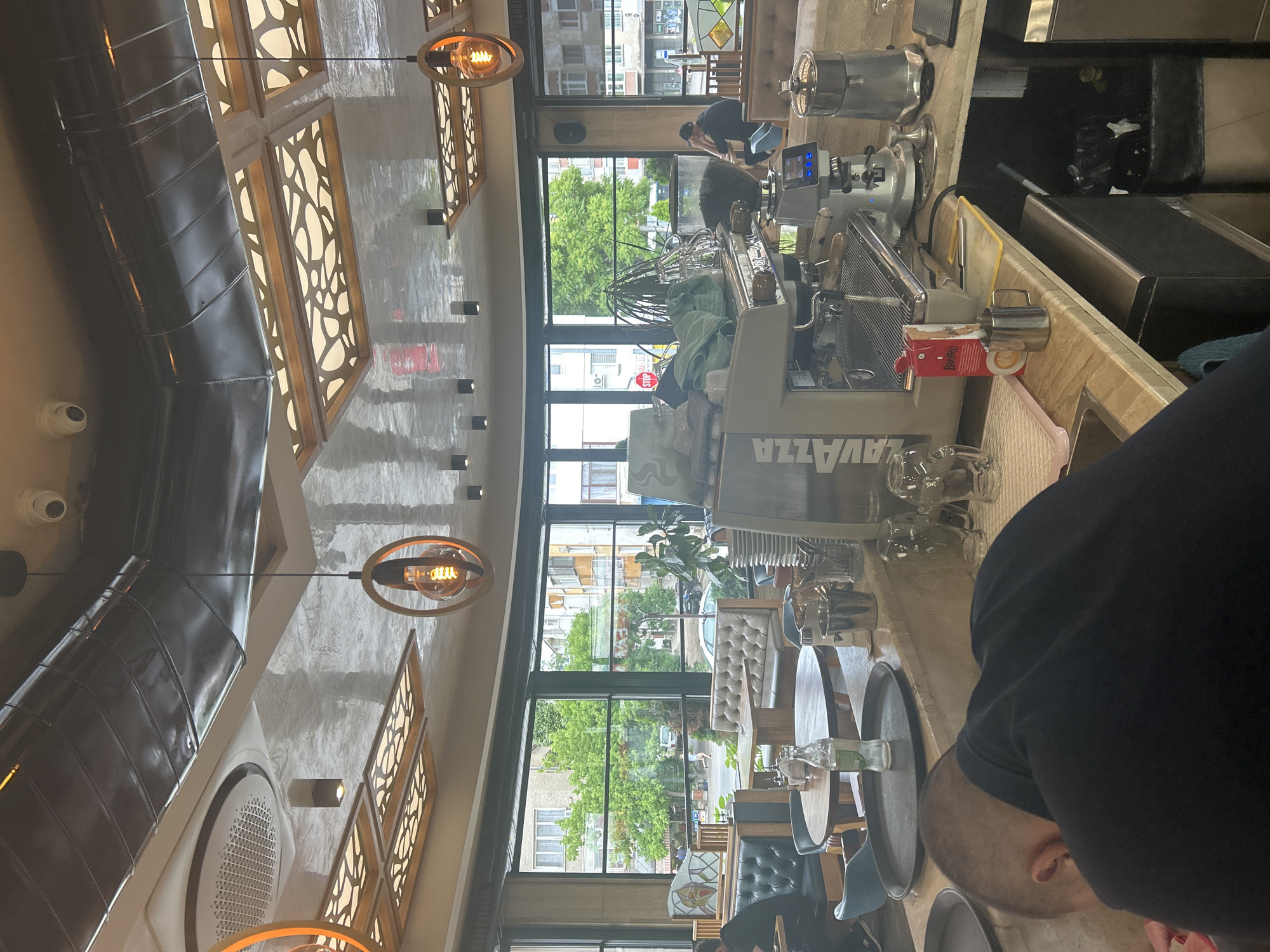 The interior of a cafe or restaurant in Kazanlak, Bulgaria is shown in the image. A man in a black shirt is facing the camera, partially obstructing the view of the countertop. A Lavazza espresso machine is visible, along with various glassware and serving trays. Windows provide a view of the outside, revealing a street scene with buildings and trees. Inside the cafe, tables and chairs are arranged near the windows, where people can be seen sitting. The ceiling is decorated with patterned panels and modern light fixtures. Air conditioning vents and security cameras are attached to the ceiling.