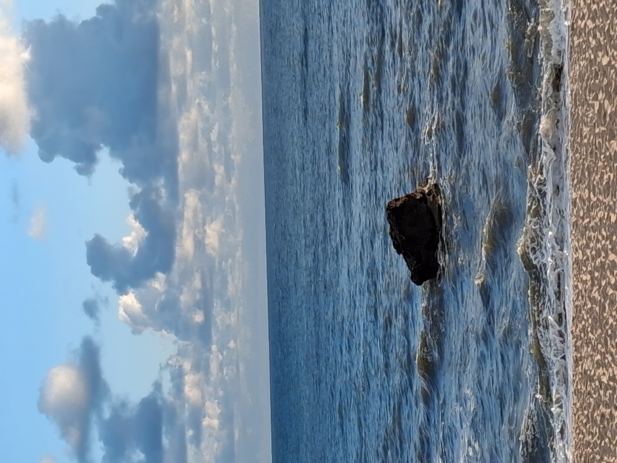 The image depicts a coastal scene likely taken in Figueira da Foz, Portugal, given the characteristics of its beaches and coastline. The scene features a body of water, probably the Atlantic Ocean, occupying a significant portion of the frame. The water appears turbulent, with visible waves and textures. A large dark rock is positioned within the water, close to the shore. The shoreline itself is composed of sand or pebbles. The sky is filled with clouds, adding depth and interest to the scene. The clouds vary in color and density, suggesting changing weather conditions.