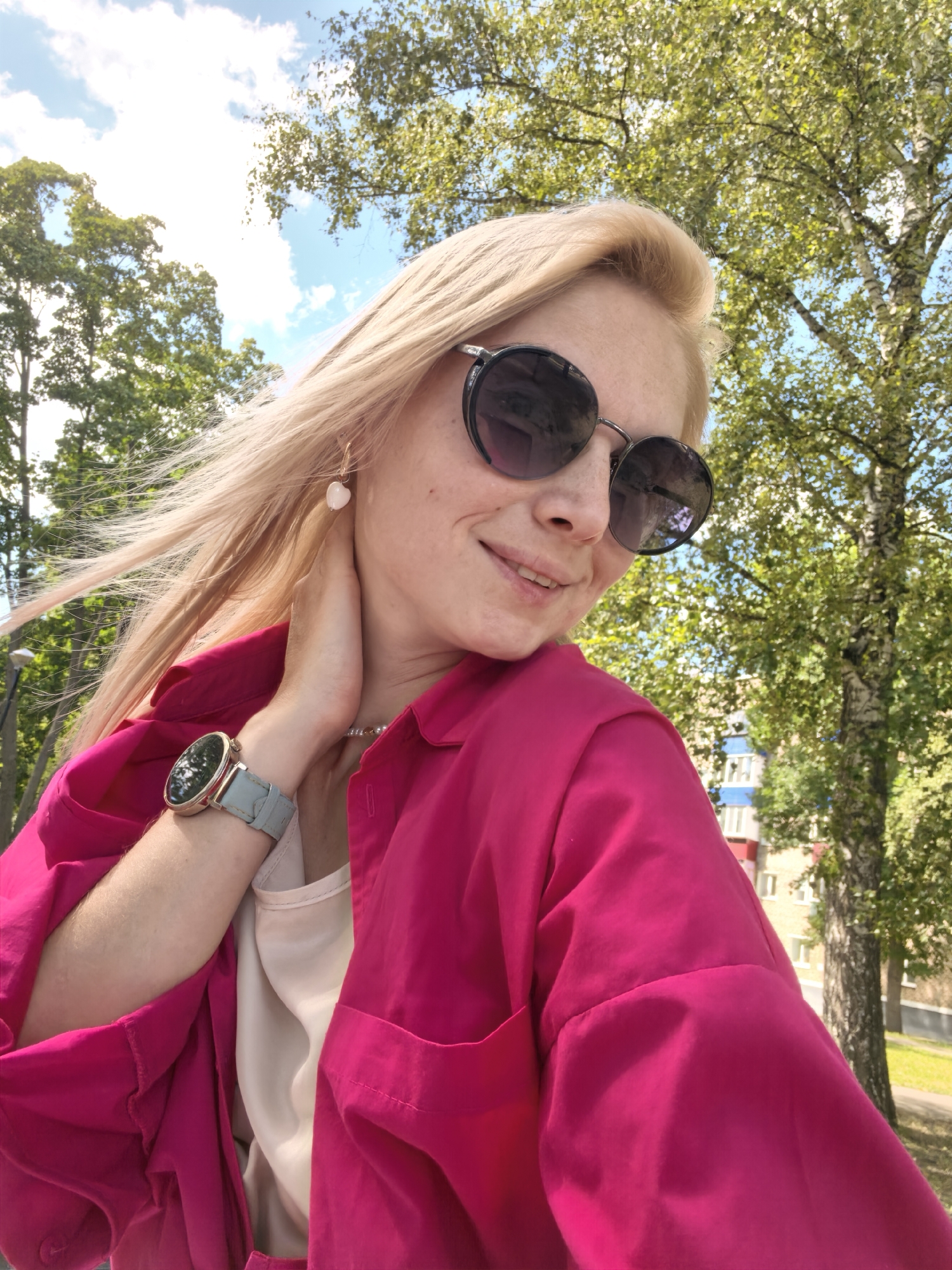 A woman with blonde hair and sunglasses is taking a selfie outdoors. She is wearing a bright pink jacket over a cream-colored top and a watch with a grey strap. Her left hand is touching her neck, and her right arm is extended to hold the camera. The background shows trees with green leaves and a building. The sky is blue with scattered clouds. The image is taken during the daytime. No specific landmarks or details indicating the city of Sterlitamak, Russia, are visible.