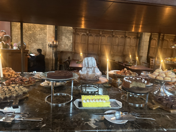 The image displays an indoor dessert buffet within a large, historic room located in Cernay-la-Ville, France. The room's background features stone block walls and sections of dark wooden paneling with carved details. The lighting is predominantly warm and dim, emanating from multiple lit taper candles in silver holders on the buffet and candelabras in the background, creating a historic atmosphere.
In the foreground, a dark, polished marble-effect countertop serves as the buffet table. It is laden with various desserts: a round dark chocolate cake on a silver stand, a tall conical fluted cake dusted with powdered sugar on a wooden board, a light-colored cake with white frosting and floral decorations (partially sliced) on another silver stand, and a tiered silver stand holding multiple sugar-dusted round pastries (donuts). Other items include a rectangular yellow cake with meringue swirls on a white platter, numerous small pastries, muffins, and tarts arranged on wooden boards and platters. Silver serving tongs and spatulas are provided on small white plates.
In the left background, two individuals are partially visible near a dark wooden counter; one male figure in a vest and shirt interacts with another. Further into the mid-ground, a person dressed in dark attire stands with their back to the viewer, operating a machine or device on a stand. The deeper background reveals additional dark wooden tables and chairs, with one table draped in a dark cloth, suggesting an event or dining area.