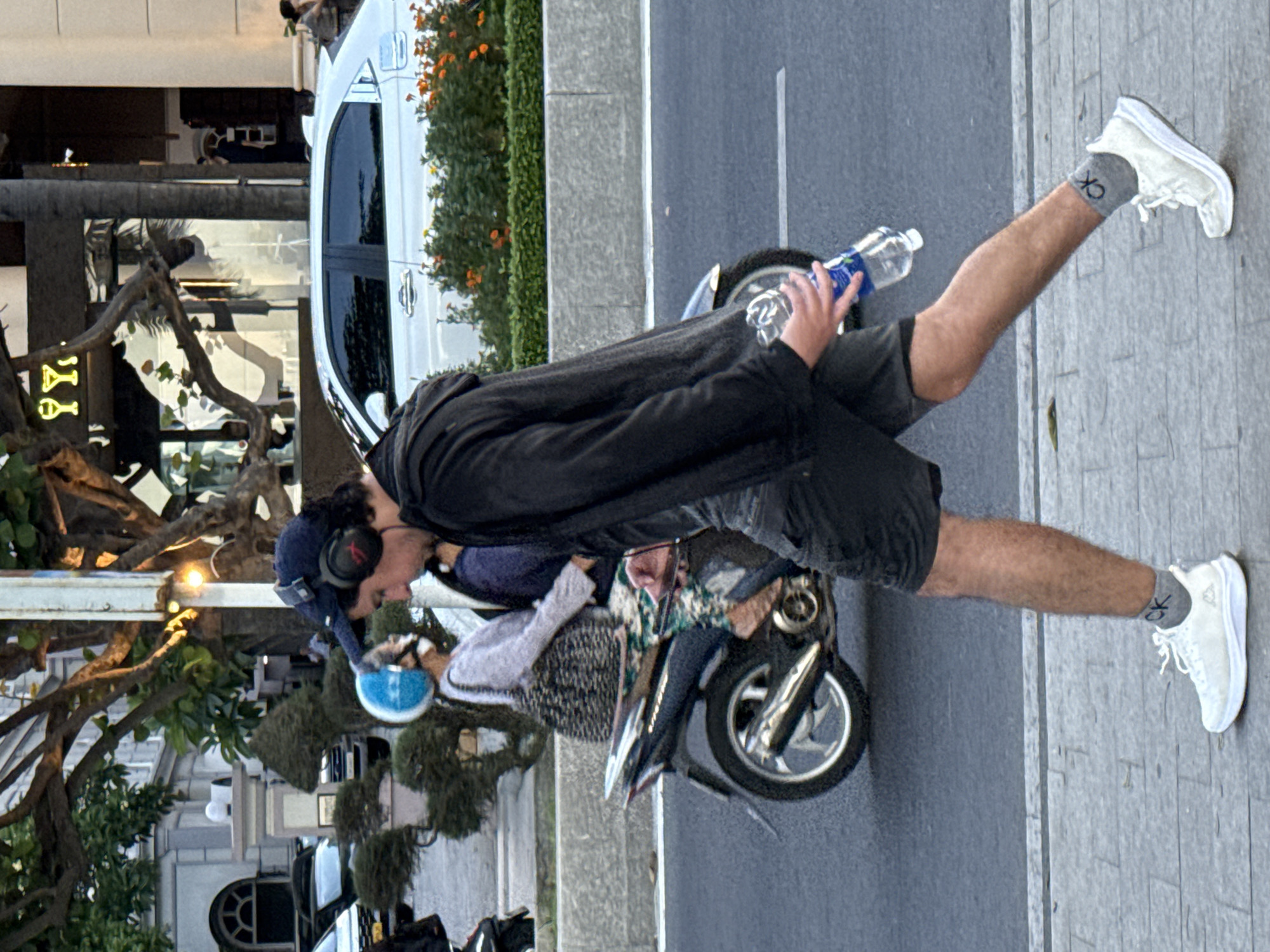 A man wearing a black hoodie, black shorts, white athletic shoes, and grey socks with "CK" logos is walking on a sidewalk in Nha Trang Ward, Vietnam. He has headphones on and is holding a clear plastic water bottle with a blue label. To his left, a woman wearing a blue helmet, a light-colored sweater, and patterned pants is riding a scooter. A white sedan is parked on the street behind the scooter, and a row of green bushes with orange flowers is visible. The background shows a building with arched windows and trees.