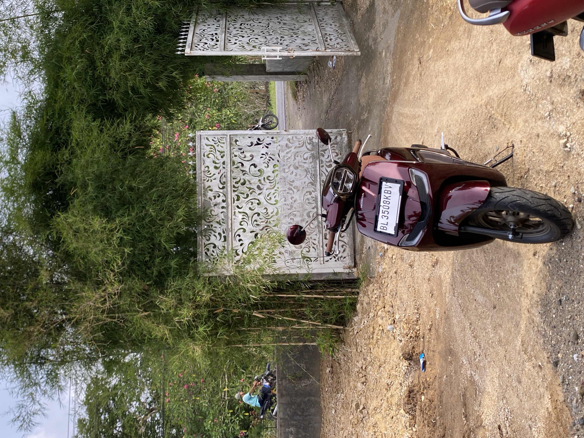 In Cottrueng, Indonesia, a maroon Honda electric scooter is parked on a dirt path in front of an open, ornate white metal gate. The gate is flanked by dense green bamboo foliage, which forms an archway over the entrance. In the background, beyond the gate, pink flowers are visible, and a person on a motorcycle is seen on a paved road. The ground is covered with loose dirt, stones, and some scattered debris. Another red scooter is partially visible in the lower right corner of the image. The sky is overcast.