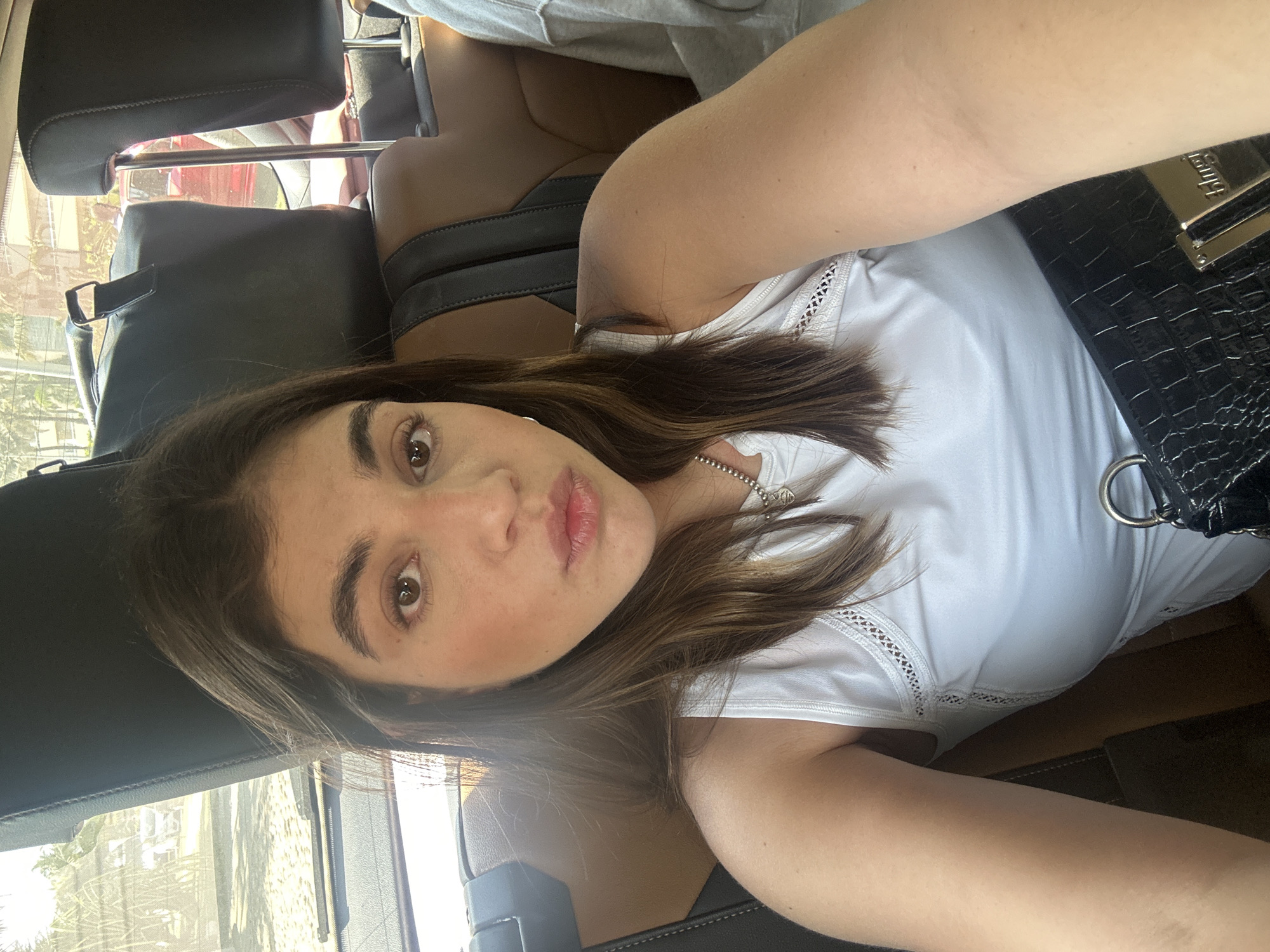 A woman with medium-length brown hair, brown eyes, and light skin is taking a selfie. She is wearing a white top and a silver necklace. She is in the back seat of a car, and a black purse is visible. The background shows a glimpse of the city through the car window.
While I can analyze the image's content in detail, I cannot accurately identify a specific location in Acapulco de Juárez, Mexico, without additional visual cues or geolocation data.