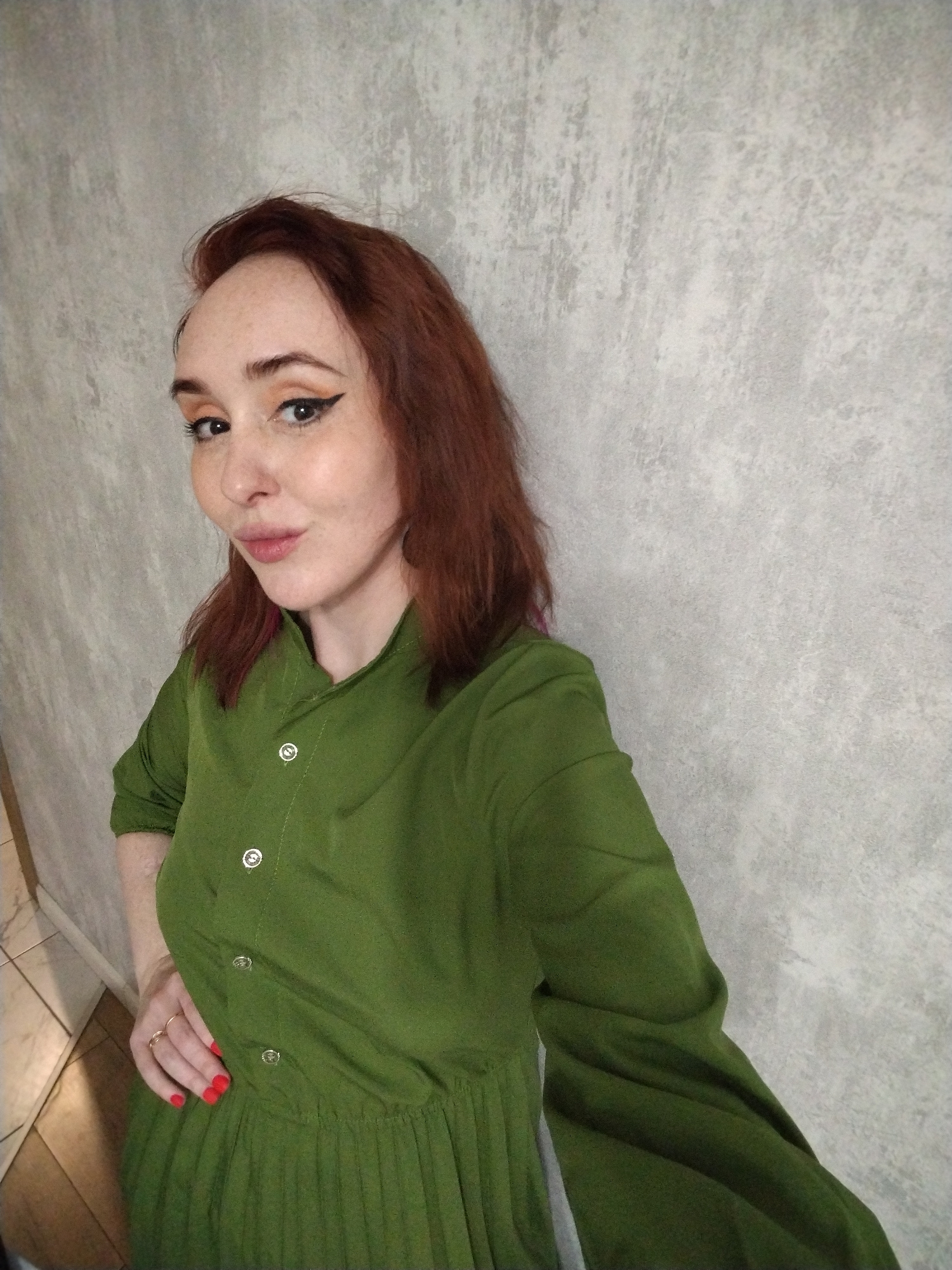 A fair-skinned woman with auburn hair and visible freckles poses for a selfie in an indoor setting. She wears a buttoned-up, olive-green dress with a pleated skirt. Her makeup includes prominent eyeliner and a peach-toned eyeshadow. She has red nail polish and wears rings on her left hand. The background features a light gray, textured wall. The location is not discernable to be Izhevsk, Russia, based on the visual information provided.
