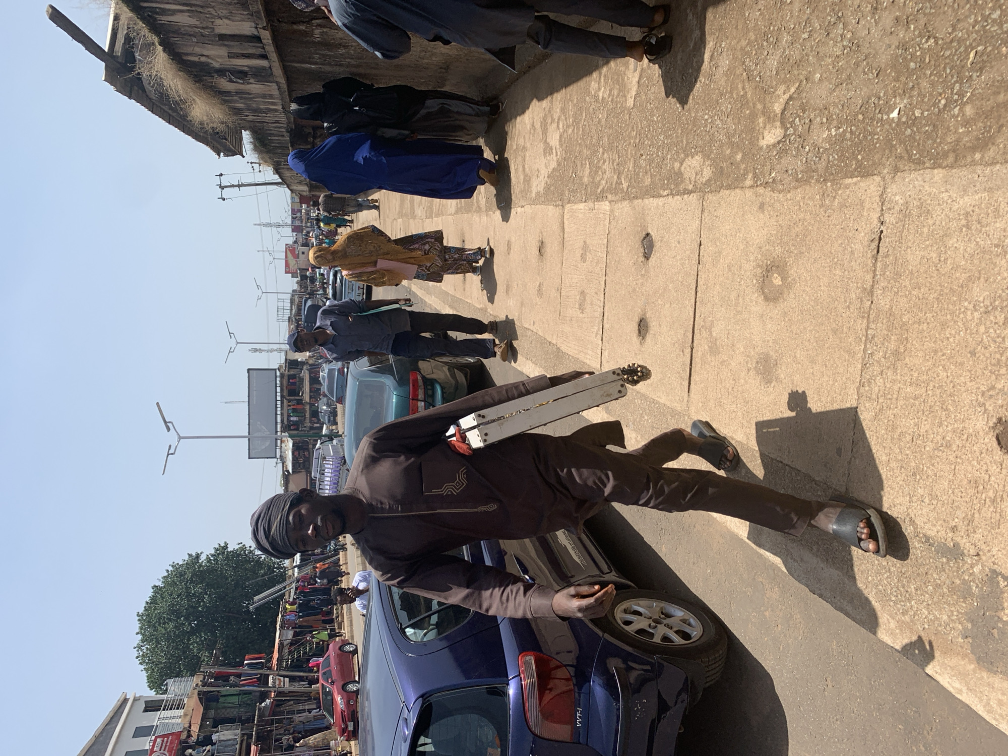 A man in a brown traditional outfit and headwrap walks on a concrete sidewalk in Jos, Nigeria, carrying a silver and white case. He is on the right side of the image, walking towards the viewer's left. The sidewalk is bordered by a street with vehicles and a bustling market. A blue car is parked on the left side of the street, and other cars and motorcycles are visible further down. The market appears to have stalls set up along the street, with various goods on display. Several other pedestrians are walking on the sidewalk and in the street. The environment is sunny and urban, with a mix of concrete structures, some in disrepair, and vegetation.