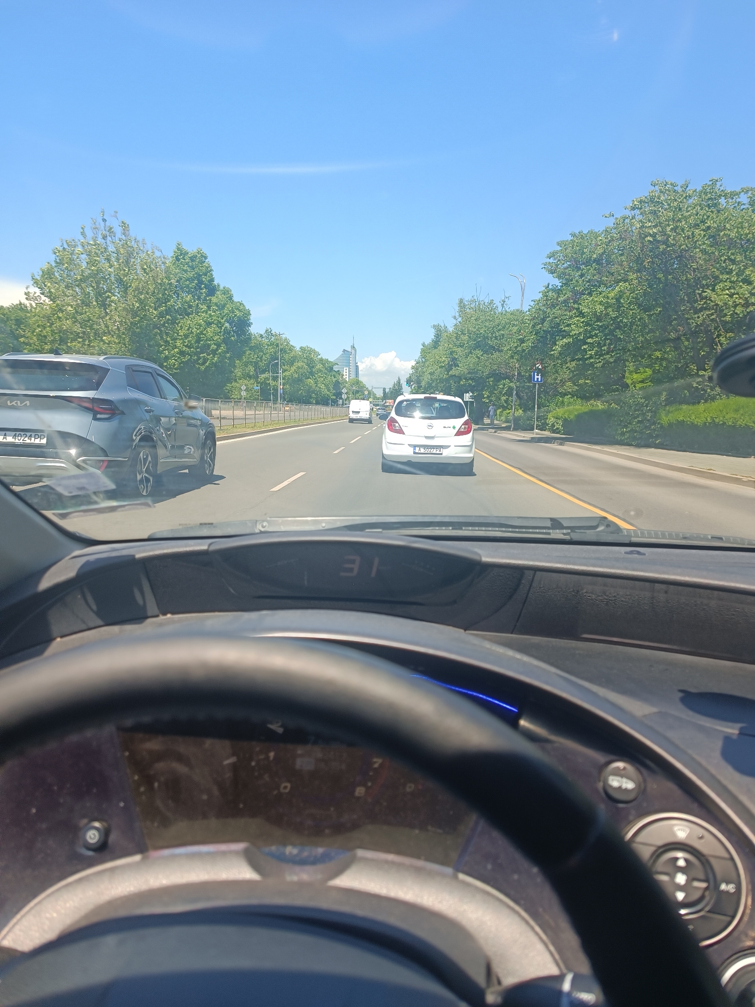 The image is a first-person perspective from inside a car, looking forward. The dashboard and steering wheel are visible in the foreground. The car is driving on a multi-lane road with a median and trees lining the sides. Several other vehicles are on the road, including a grey SUV on the left, a white hatchback in the middle, and a white van further ahead. Buildings and a cloudy sky are visible in the distance. The car's digital display shows "31".
This scene is captured on Tsvetan Dimov Street in Burgas, Bulgaria, specifically in the vicinity of the "Izgrev" district, near the intersection with "Lazar Lydzhev" street and close to the "Panteon" monument. The grey SUV with license plate "A 4024 PP" is a Kia Sportage, and the white hatchback is a Opel Corsa with license plate "A 5027 PA". The street is bordered by lush green trees and on the right side, a bus stop sign with an 'H' indicating a bus stop is visible, and a pedestrian can be seen walking on the sidewalk.