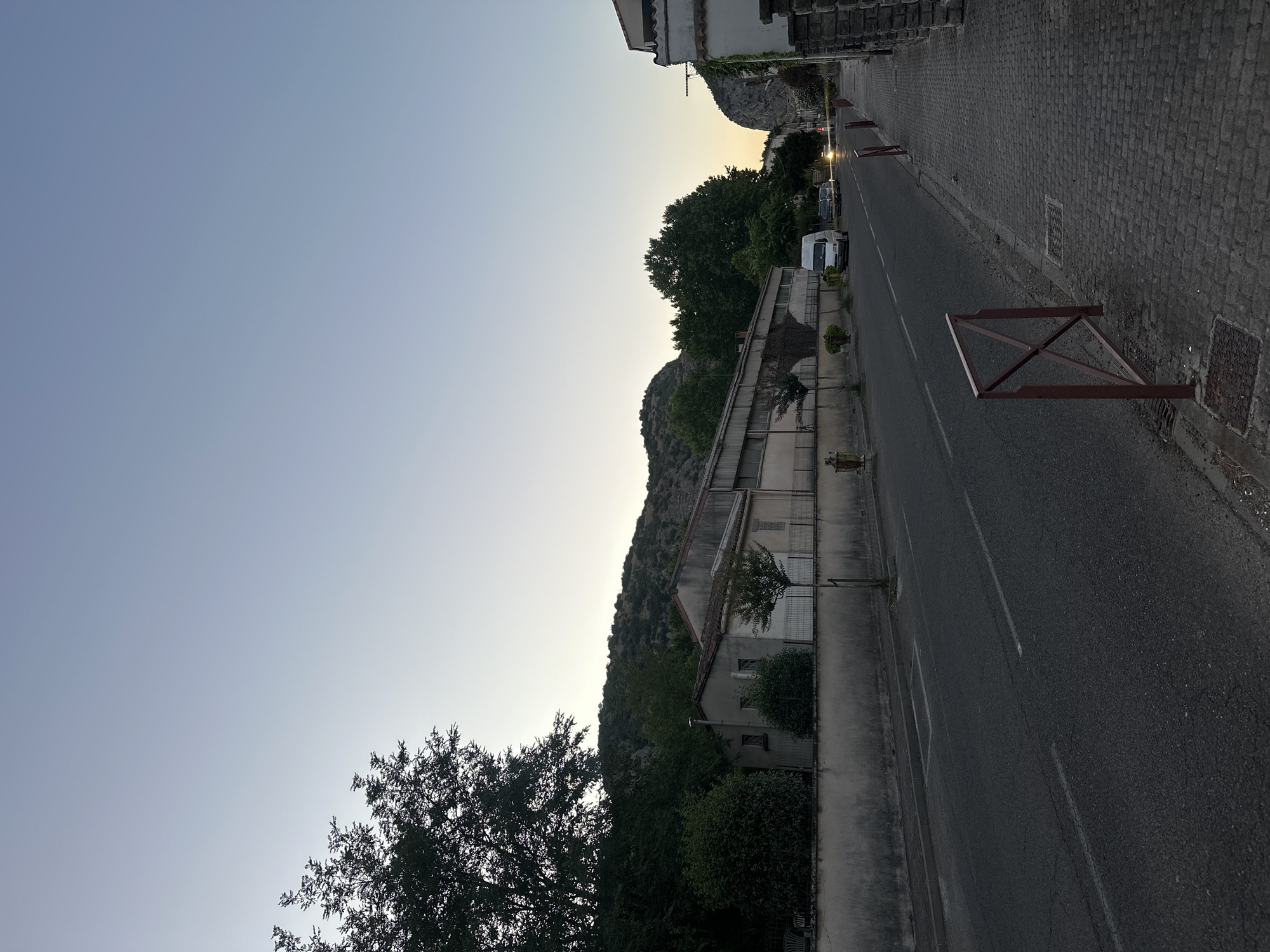 The image depicts a street scene in Anduze, France, likely during twilight. On the left, a long, low-rise building with a grey, weathered facade runs parallel to the road. It appears to be an industrial or commercial structure, with a fence running along its front. Trees and dense foliage are visible behind the building and on a rocky, tree-covered hillside in the background. The road in the foreground is asphalt, with white lane markings. A paved sidewalk runs along the right side of the road, constructed with cobblestones. Red metal railings are placed intermittently along the edge of the sidewalk. Several vehicles are present on the road, including a white van and two dark cars. The sky is a clear, pale blue, transitioning to a lighter hue towards the horizon, suggesting the setting or rising sun. The overall impression is one of a quiet, somewhat dated urban environment bathed in soft, natural light.