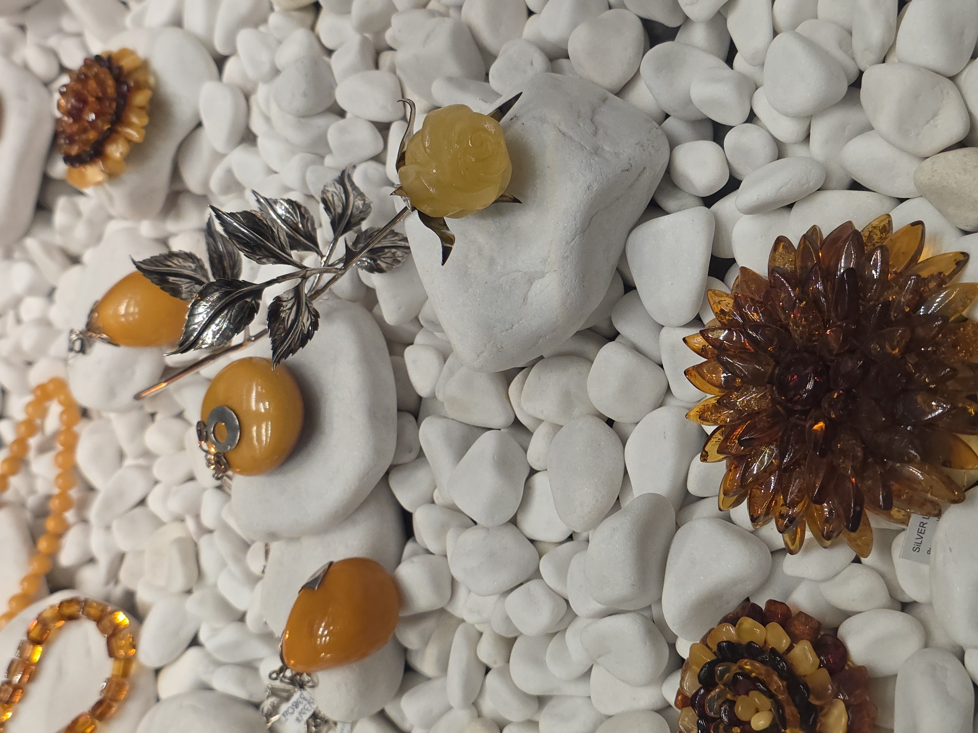 The image shows an assortment of amber jewelry, including flower-shaped brooches, pendants, and a necklace, displayed on a bed of small, white pebbles. One pendant has a silver branch attached. The brooches feature varying shades of amber, from dark brown to honey-colored. Location data, if any, is not discernible from the image. The city of Warsaw, Poland is irrelevant without further data.