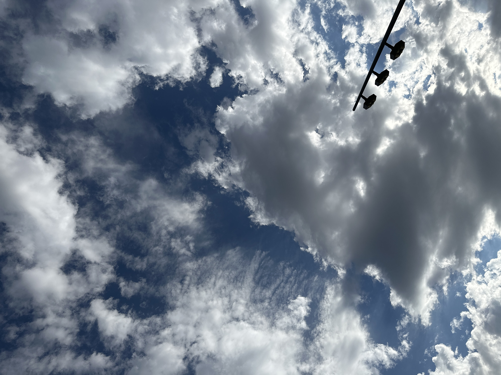 The image captures a dynamic sky with a mix of white and dark gray clouds against a deep blue sky. In the lower right corner, a black pole with three light fixtures is silhouetted against the clouds. The lighting suggests it is daytime, possibly late afternoon or early morning, given the dramatic cloud formations and the angle of light. The scene appears to be an outdoor urban setting in Tashkent, Uzbekistan, possibly a park or public square, indicated by the presence of the street light. There are no people or discernible actions depicted.