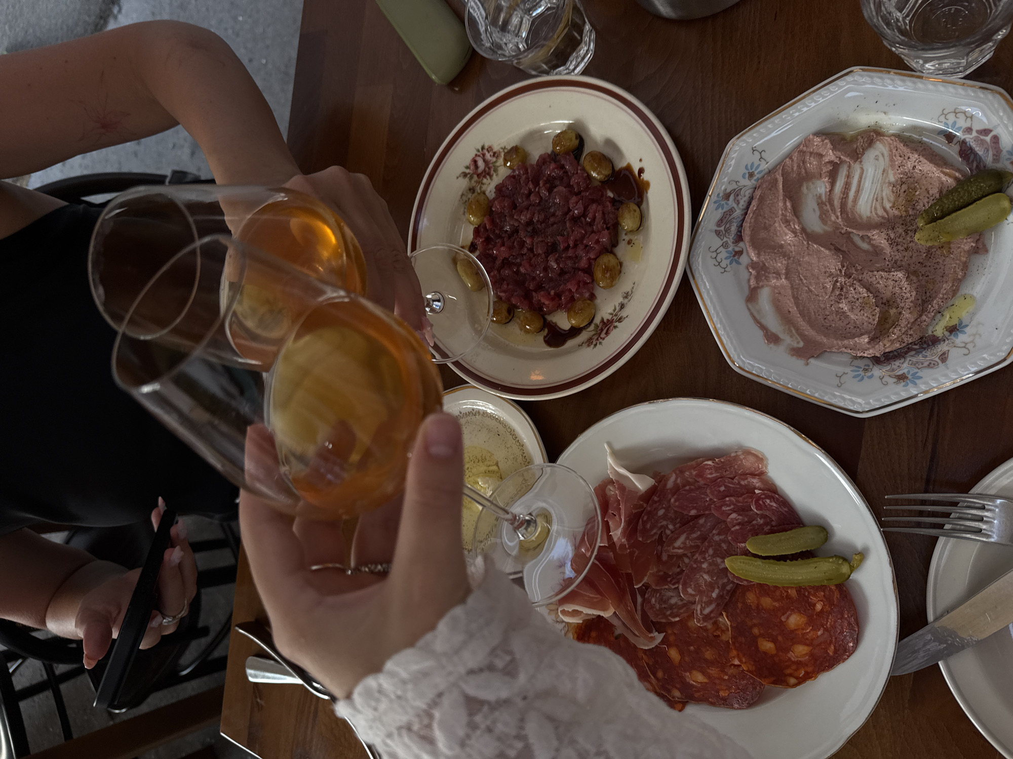 The image depicts a scene on a wooden table, likely in a restaurant in Belgrade, Serbia. Two glasses of amber-colored liquid, possibly wine or aperitif, are held by unseen individuals, one raised as if in a toast. The glasses are set against a backdrop of various food dishes.
In the foreground, a white plate holds an assortment of cured meats, including slices of chorizo and prosciutto, accompanied by two cornichons. To the right of this plate, another white, octagonal plate presents a pinkish spread, possibly a dip or pâté, swirled with lighter cream-colored streaks and garnished with two cornichons and a small dollop of yellow condiment.
Further into the frame, a circular plate with a floral rim features a dish of what appears to be steak tartare, a mound of finely chopped raw beef, surrounded by several small, golden-brown spheres, possibly roasted grapes or potatoes. A small glass of water with ice is also visible on the right side of the table.
In the upper left quadrant, one person's hand is holding a smartphone. The environment appears to be indoors, with the presence of cutlery and the overall arrangement suggesting a dining setting. The lighting is warm, highlighting the textures and colors of the food and drinks.