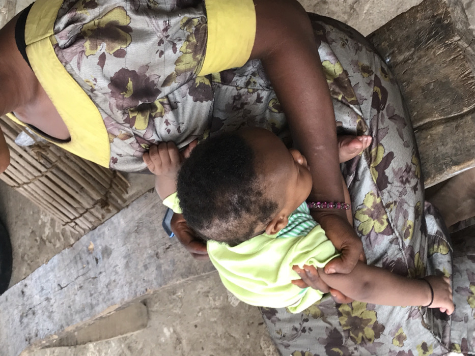 Overhead shot of a woman holding a baby in her lap. The woman wears a dress with a floral pattern and yellow trim, along with a beaded bracelet. The baby is dressed in a light green shirt. They sit on a weathered wooden surface, possibly a bench, with a cylindrical wicker item nearby. The ground is a light, earthy tone. The focus is on the intimate moment between the woman and child.