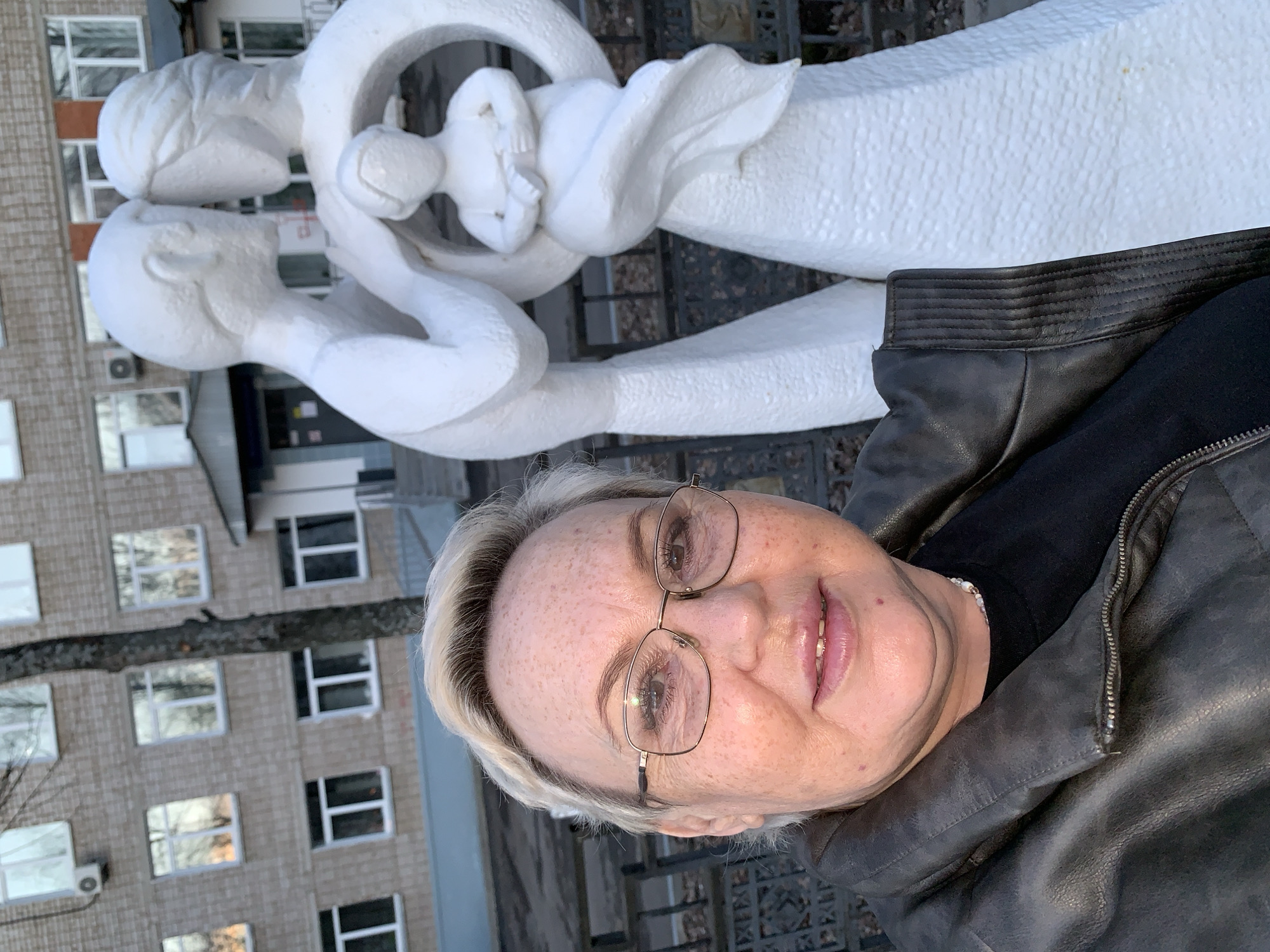 The image captures a woman posing in Chaikovsky, Russia, with an abstract sculpture and a building in the background. The woman, shown in a close-up, has light blonde hair, wears glasses with thin frames, and is dressed in a black leather jacket over a black top, with a subtle pearl-like bracelet on her wrist. She presents a pleasant, slightly smiling expression.
Behind her, a striking white sculpture depicts stylized, intertwined figures. Two larger forms appear to be embracing or connected, encompassing a smaller, possibly child-like figure or a central circular motif, conveying themes of family or unity. The sculpture has a textured, granular surface.
In the background, a multi-story building made of reddish-brown brick features numerous rectangular windows, some reflecting parts of the environment. An air conditioning unit is visible on its exterior. The scene appears to take place during the daytime, under potentially cool or overcast weather conditions, as suggested by the woman's attire. No clear text is visible on the sculpture, the woman, or the building.
