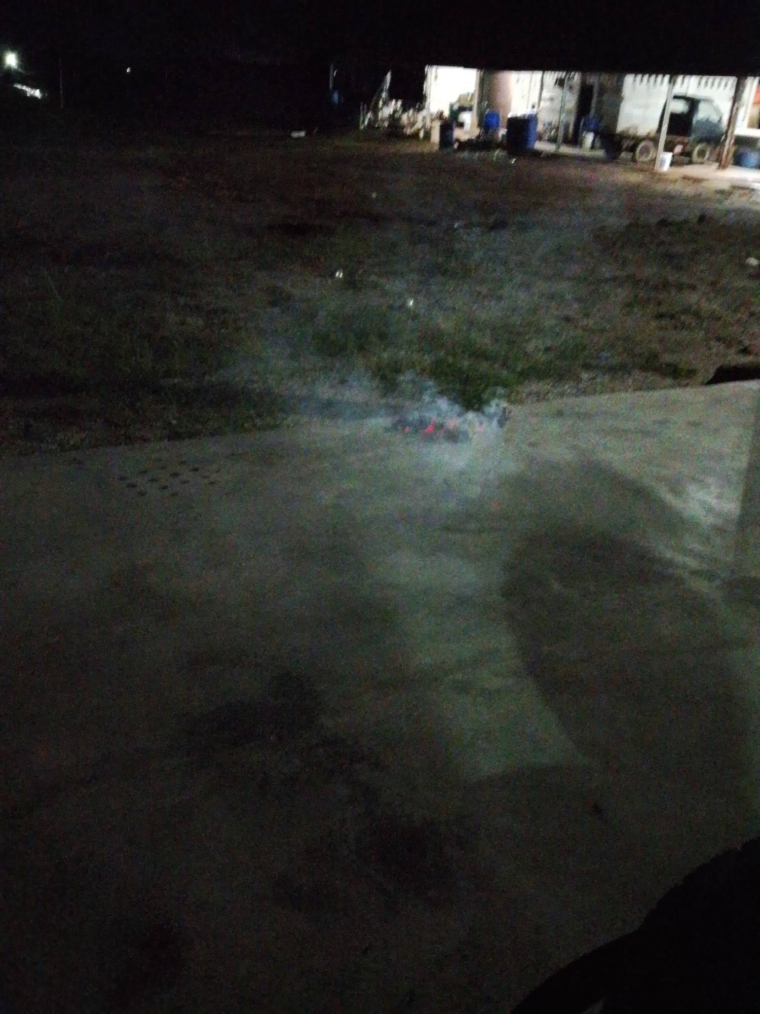 The image depicts an outdoor scene at night in Bukit Ketok, Indonesia. In the background, there is a building with an open front where various items are stored, including blue containers and what appears to be a truck or vehicle parked near the right side. The foreground is dominated by a concrete surface. In the middle ground, there is grass and some smoke rising from a small area with embers, possibly a small fire. The overall lighting is poor.