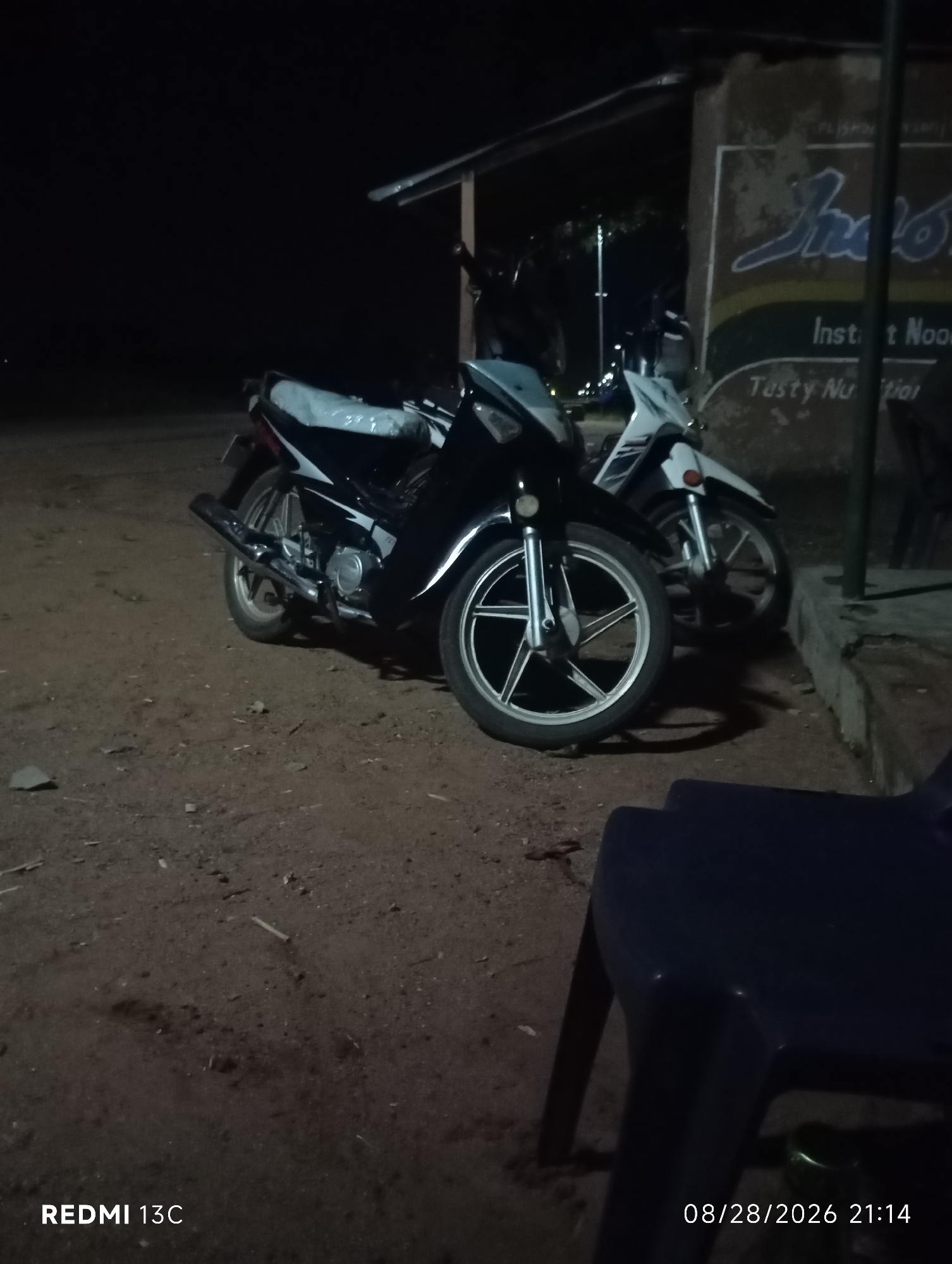 At night in Shendam, Nigeria, two motorcycles are parked on a dirt lot near a building with a sign for "Inst nt Nood" and "Tasty Nu tion." A dark blue plastic chair is in the foreground. The motorcycles are positioned side-by-side. The one on the left is black, with a white seat cover and a silver exhaust pipe. The motorcycle on the right is white and blue. The building behind them has a rustic appearance with some peeling paint. The lighting is dim, indicating it is nighttime. The ground is uneven and appears to be a mixture of dirt and small stones. A timestamp in the lower right corner reads "08/28/2026 21:14" and "REDMI 13C" is in the lower left corner.
