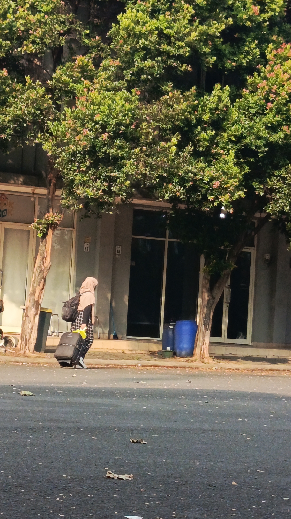 A person wearing a hijab and checkered shirt walks along a paved sidewalk beside a street in Tangerang, Indonesia. The individual is pulling a black suitcase and carrying a black backpack. They are walking past a building with large windows and trees lining the sidewalk. Two large blue barrels are visible near the building's entrance. The street is paved and has fallen leaves scattered across it. The scene is illuminated by natural sunlight.