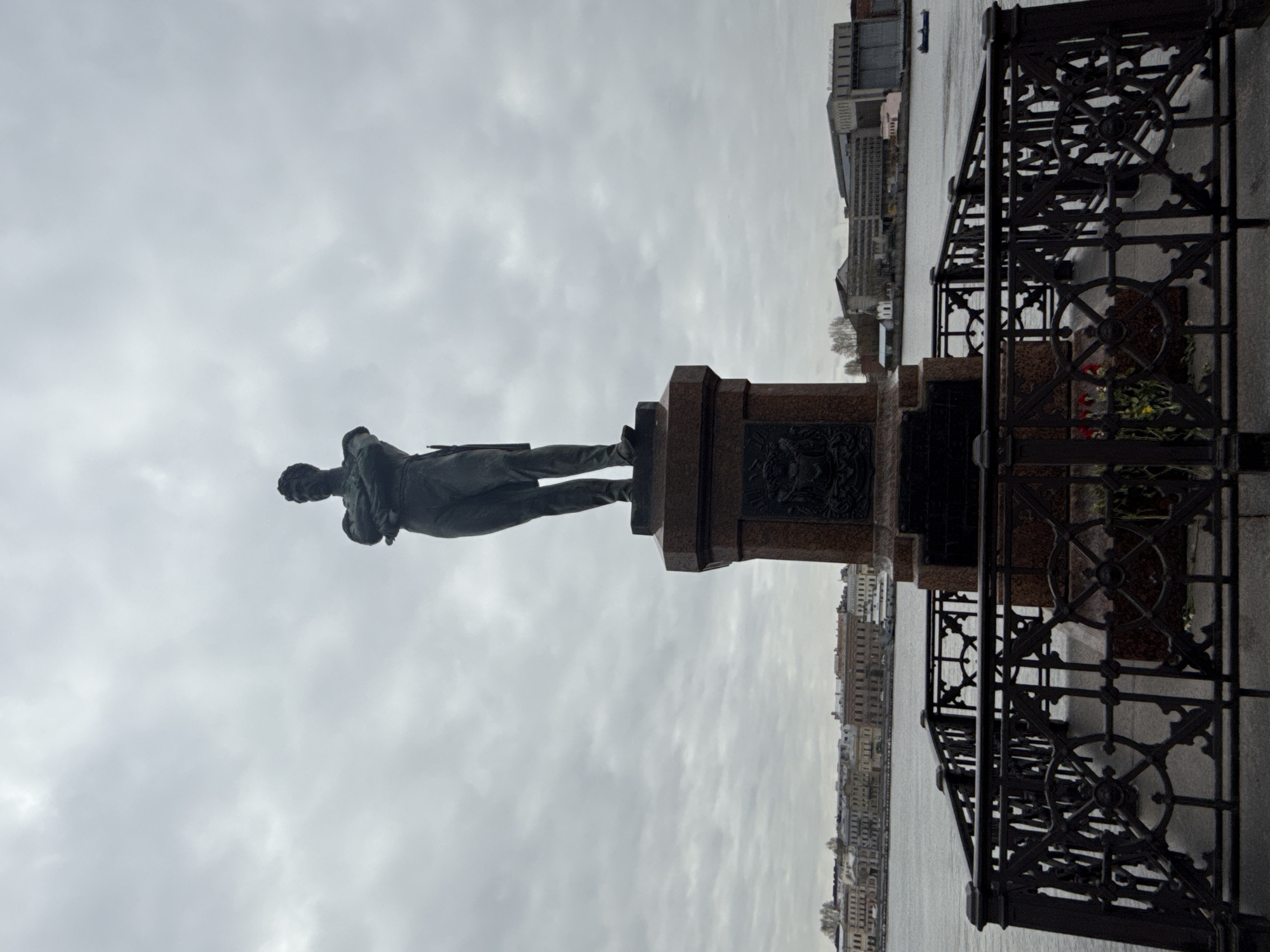 The image displays a large, dark-colored statue of a man standing on a tall, multi-tiered pedestal made of reddish-brown stone, likely granite. The statue depicts a historical figure dressed in formal attire, possibly a coat or uniform. The visible side of the pedestal features indistinct bas-reliefs or plaques.
The statue is situated on an embankment alongside a wide body of water, which appears to be a river or canal, characteristic of Saint Petersburg, Russia. Across the water, a cityscape unfolds, showcasing a continuous line of historic, multi-story buildings on the distant bank.
In the foreground, to the right of the statue, an ornate, dark metal railing with circular and geometric patterns runs along the edge of the embankment. At the base of this railing, near the pedestal, a small arrangement of red and yellow flowers is visible, suggesting a recent tribute or memorial.
The sky is overcast with thick, gray clouds, indicating a diffused light and a lack of direct sunlight, consistent with a cool or cloudy daytime. No specific activity is taking place beyond the monument's presence. No discernible text is visible on the statue or its pedestal. The overall scene conveys a solemn and historical atmosphere.