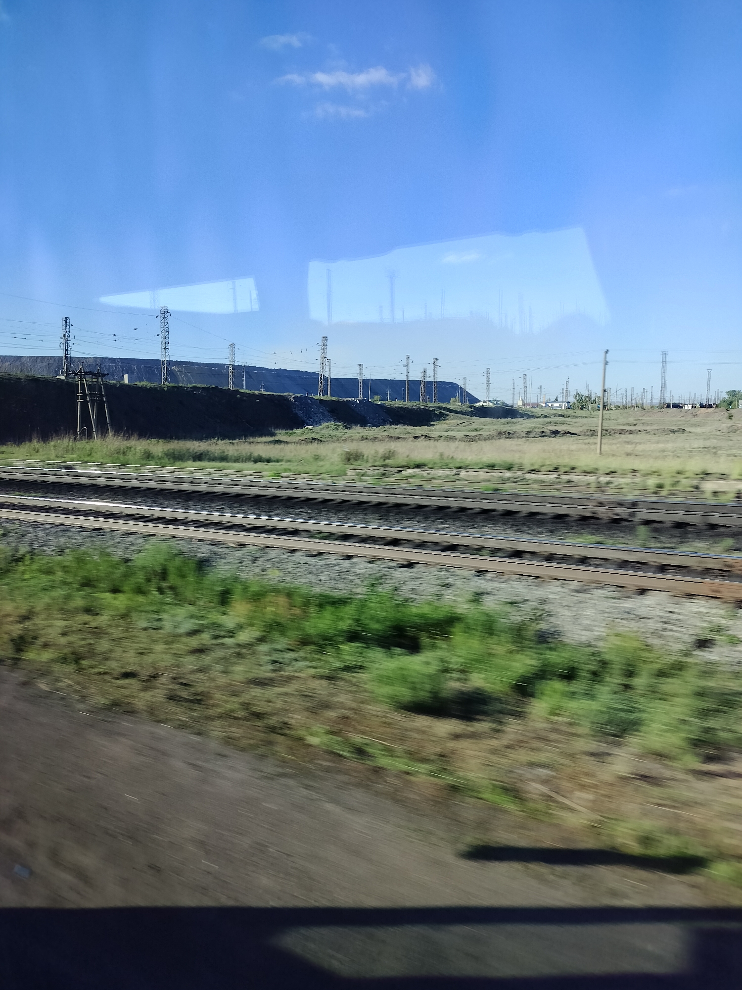 The image shows a landscape with a railroad track running through the foreground. The tracks are parallel and appear to be disused with dark ballast. Vegetation, primarily green grass and weeds, is visible on either side of the tracks. Beyond the tracks, the terrain rises slightly to form a ridge. On top of the ridge, there are numerous utility poles and power lines stretching across the horizon. In the background, the sky is blue with some light clouds. The image is likely taken from a moving vehicle, indicated by the motion blur in the foreground and the reflection in the window at the top of the frame.
The location is Unknown, Unknown. The image depicts a railroad track, a ridge with utility poles, and vegetation under a blue sky.